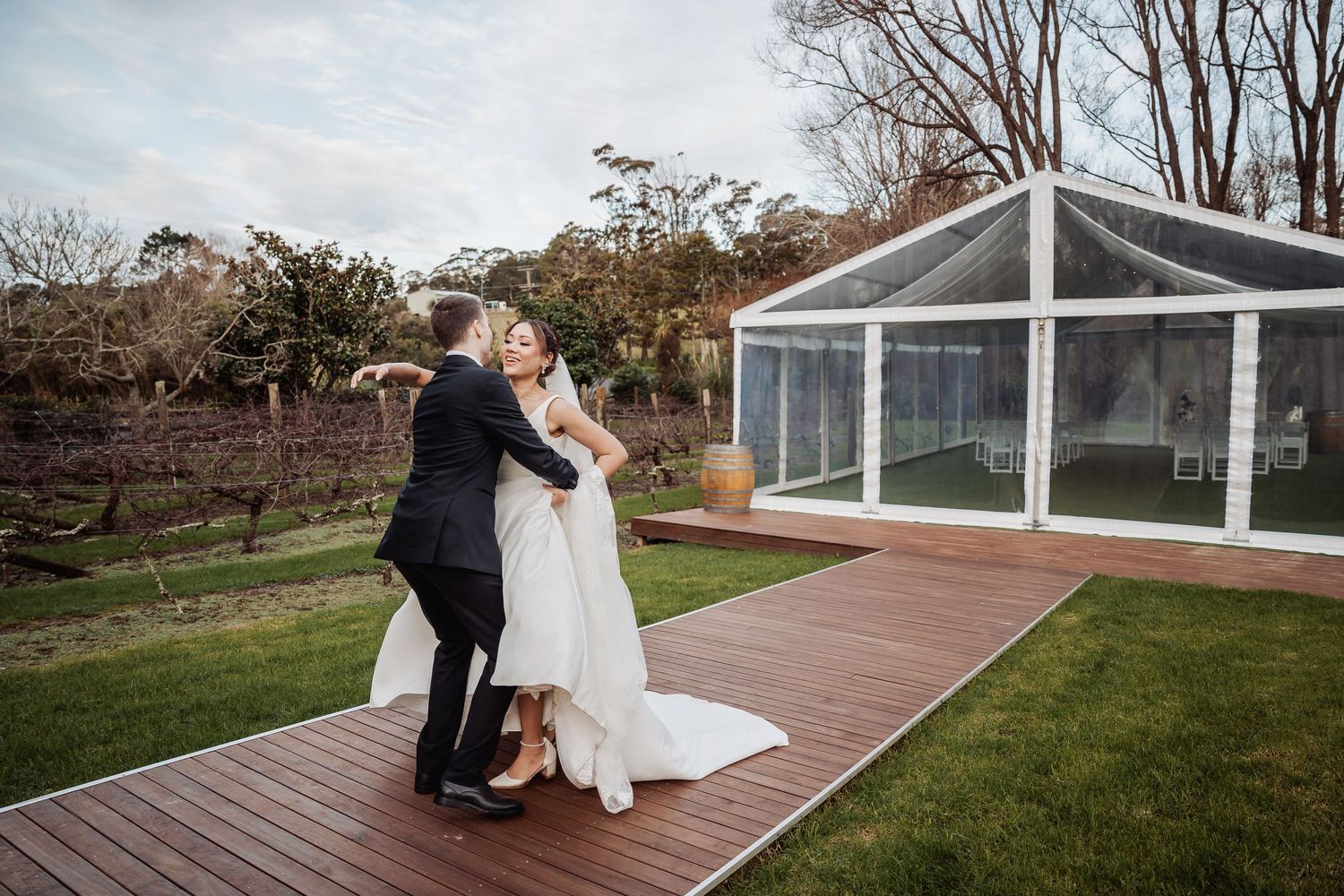 Newlyweds embrace on a wooden deck outside a modern glass conservatory building.