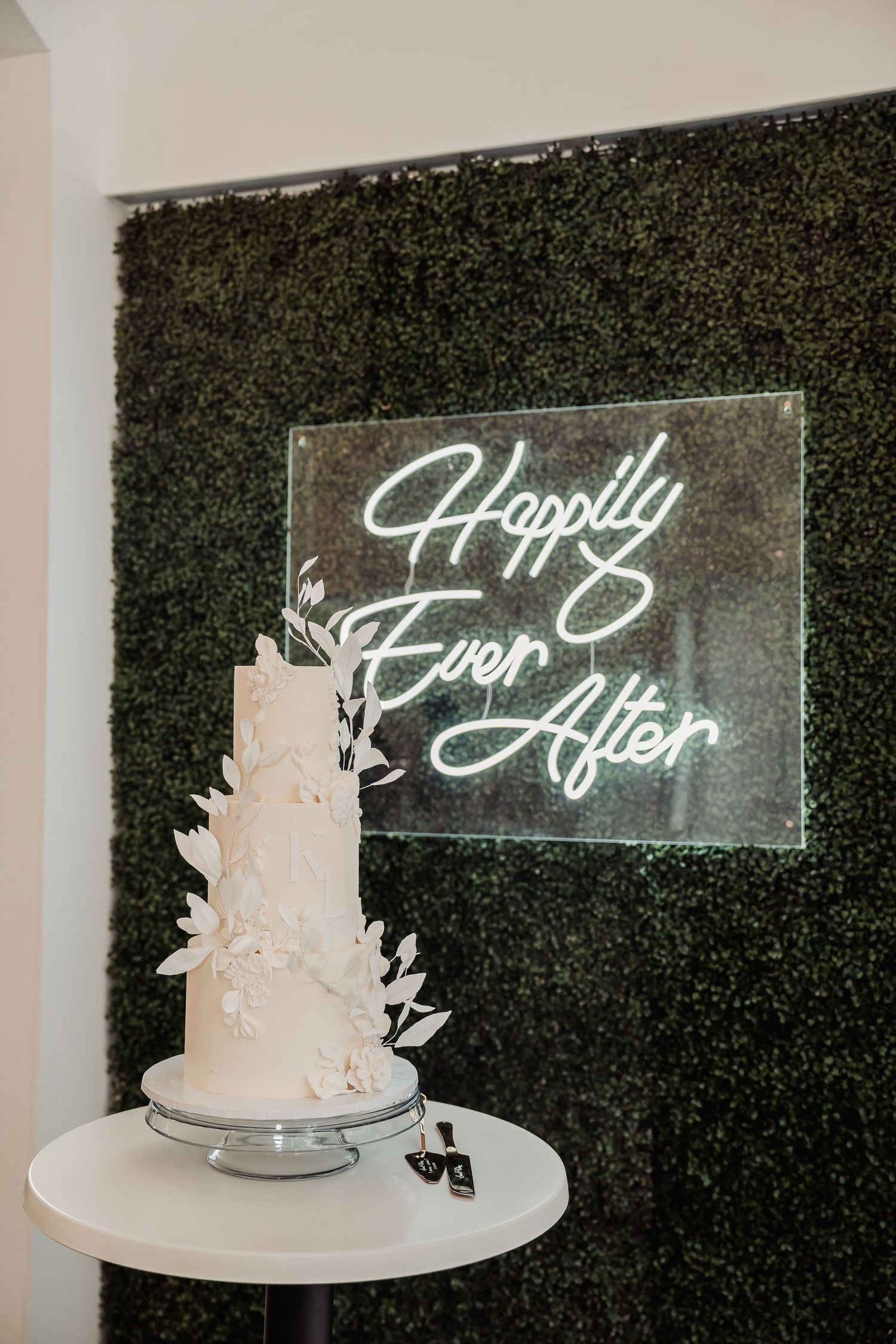 Elegant white wedding cake displayed against green hedge wall with neon 'Happily Ever After' sign.