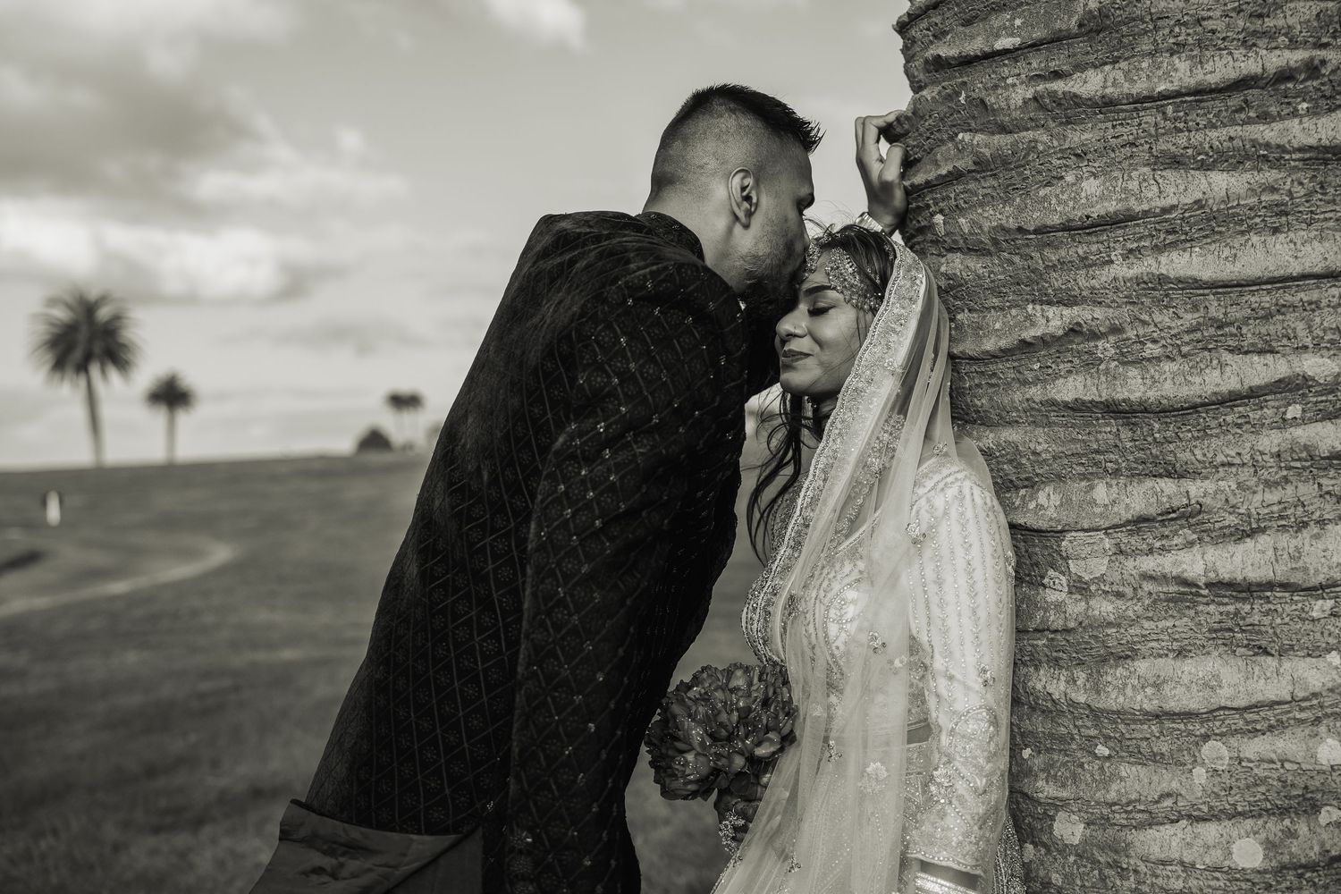 Black and white wedding portrait against palm trees showing couple in traditional attire.