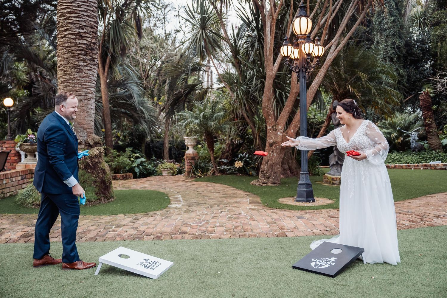 Bride and groom play cornhole at their wedding reception in a tropical garden setting.