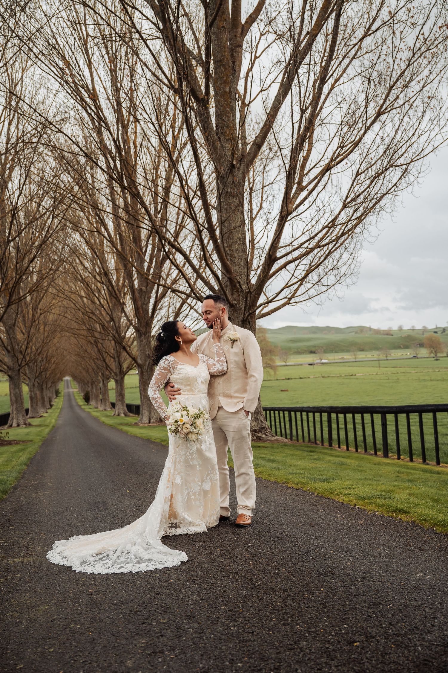 Newlyweds share a romantic moment on a tree-lined country road with rolling hills in background.