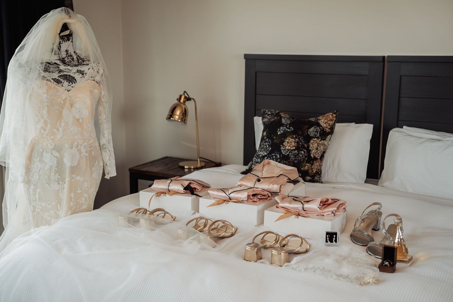 A collection of elegant wedding accessories including a white lace dress, beige heels, and jewelry laid out on a bed.