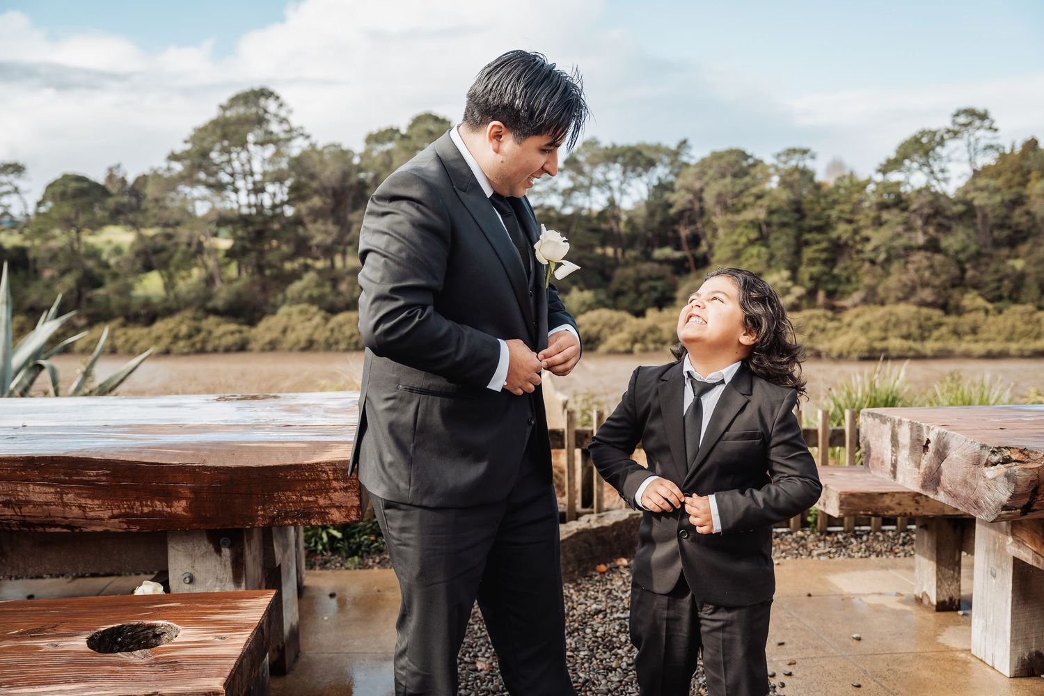 Two people in black suits sharing a lighthearted moment together on an outdoor deck near a wooded area.