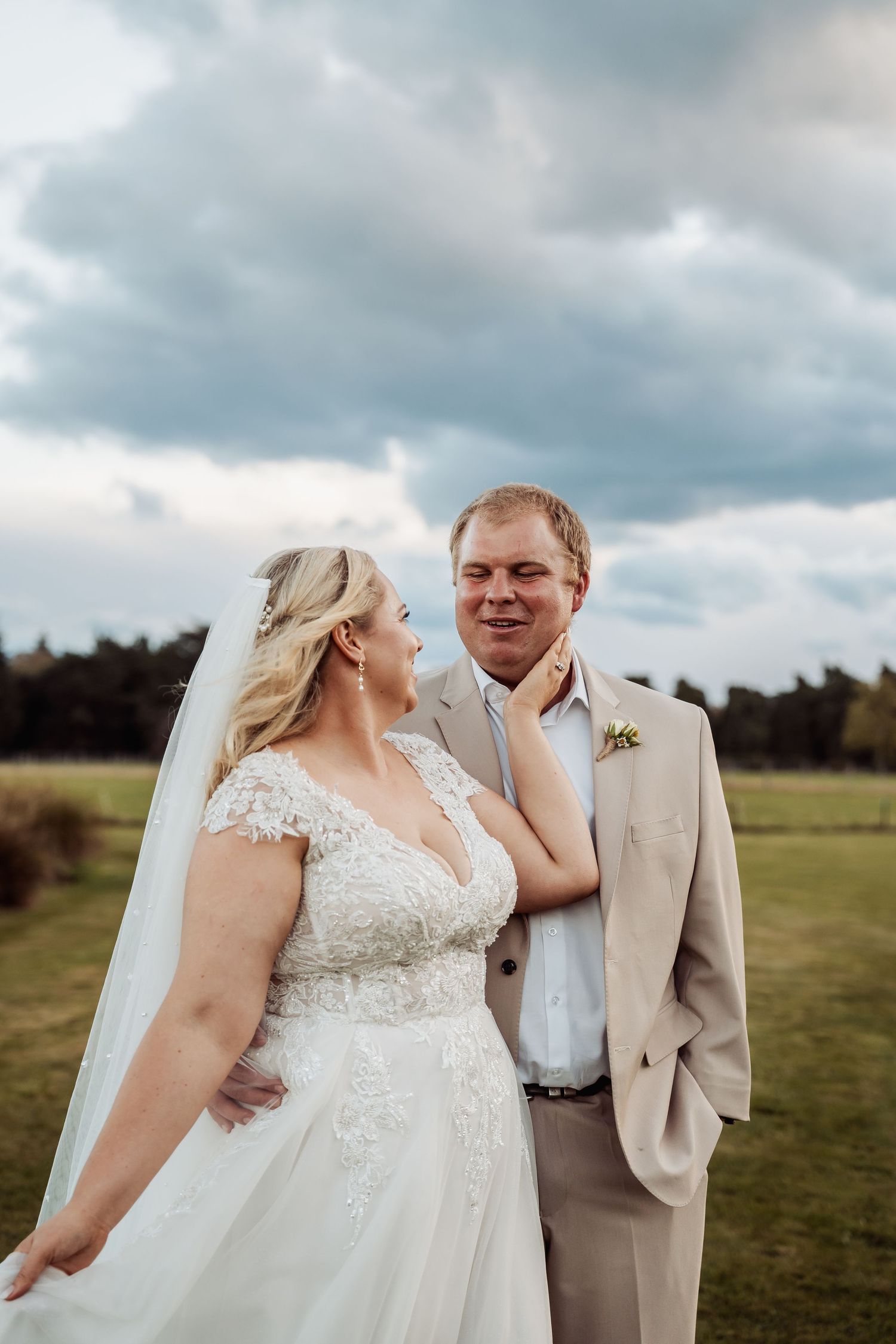 Bride and groom share tender moment in open field under dramatic cloudy sky.