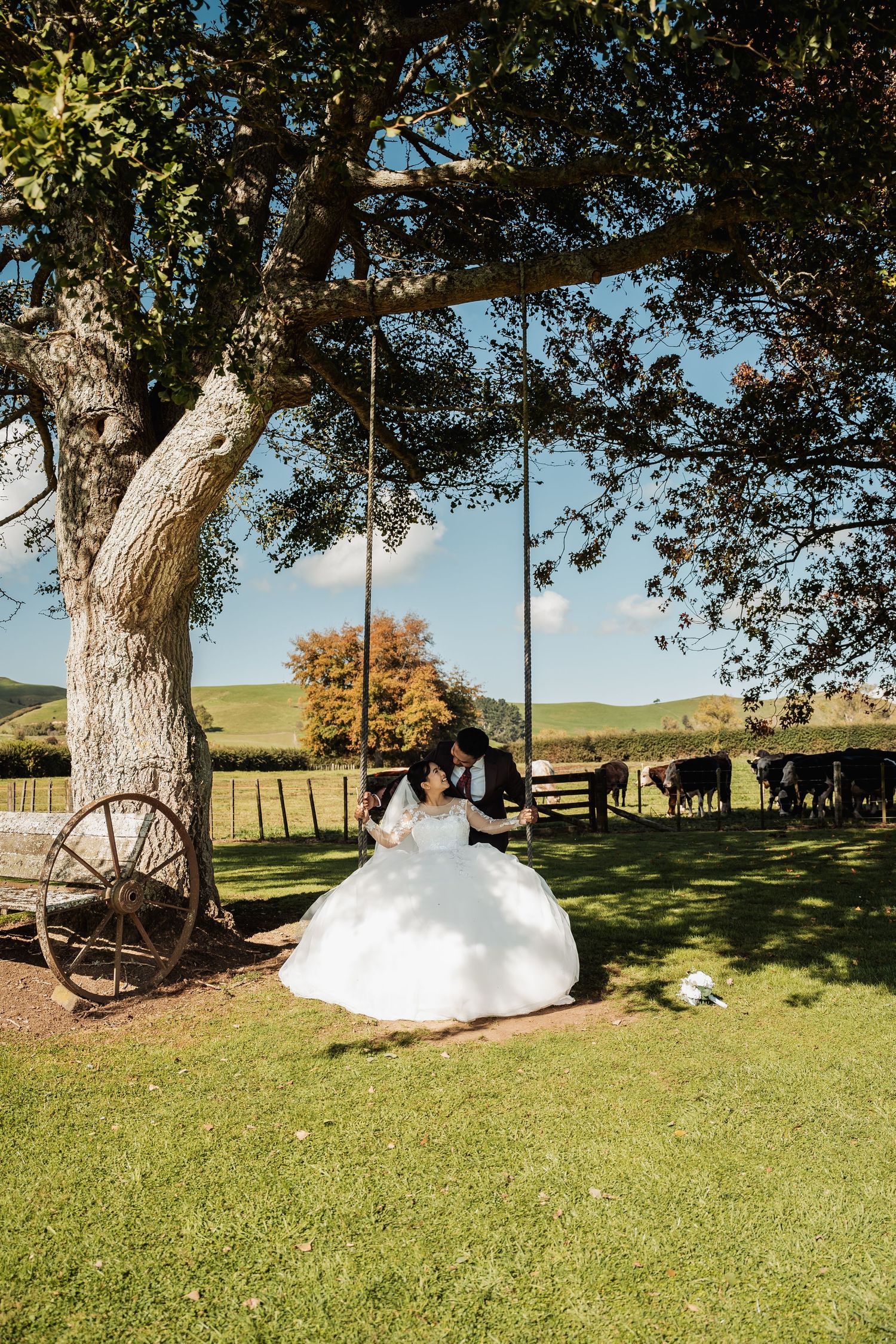 Bride and groom sit on tree swing at rustic farm wedding venue on sunny day.