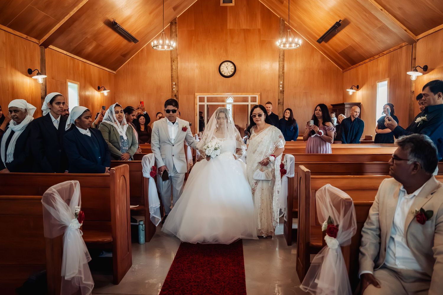 Bride walks down the aisle in a white gown at a wooden chapel wedding ceremony with guests in pews.