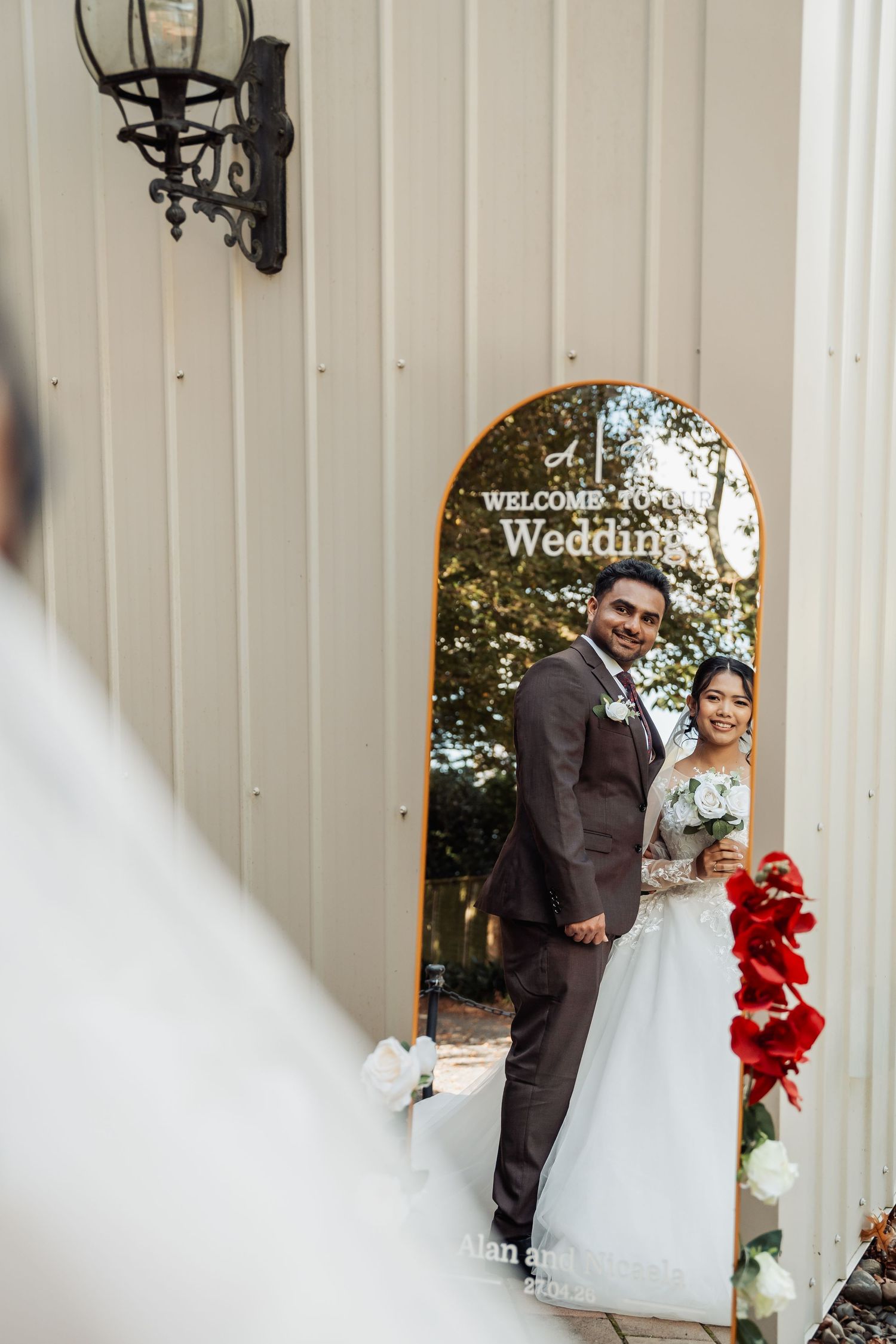 Bride and groom pose together beside a wedding welcome mirror sign at elegant white venue entrance.