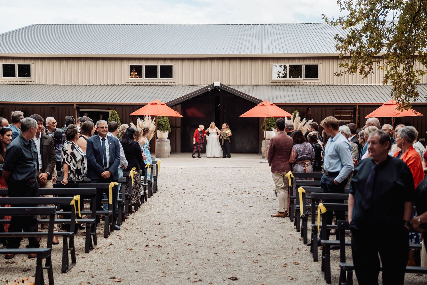 Outdoor wedding ceremony with guests seated on wooden benches at barn venue.