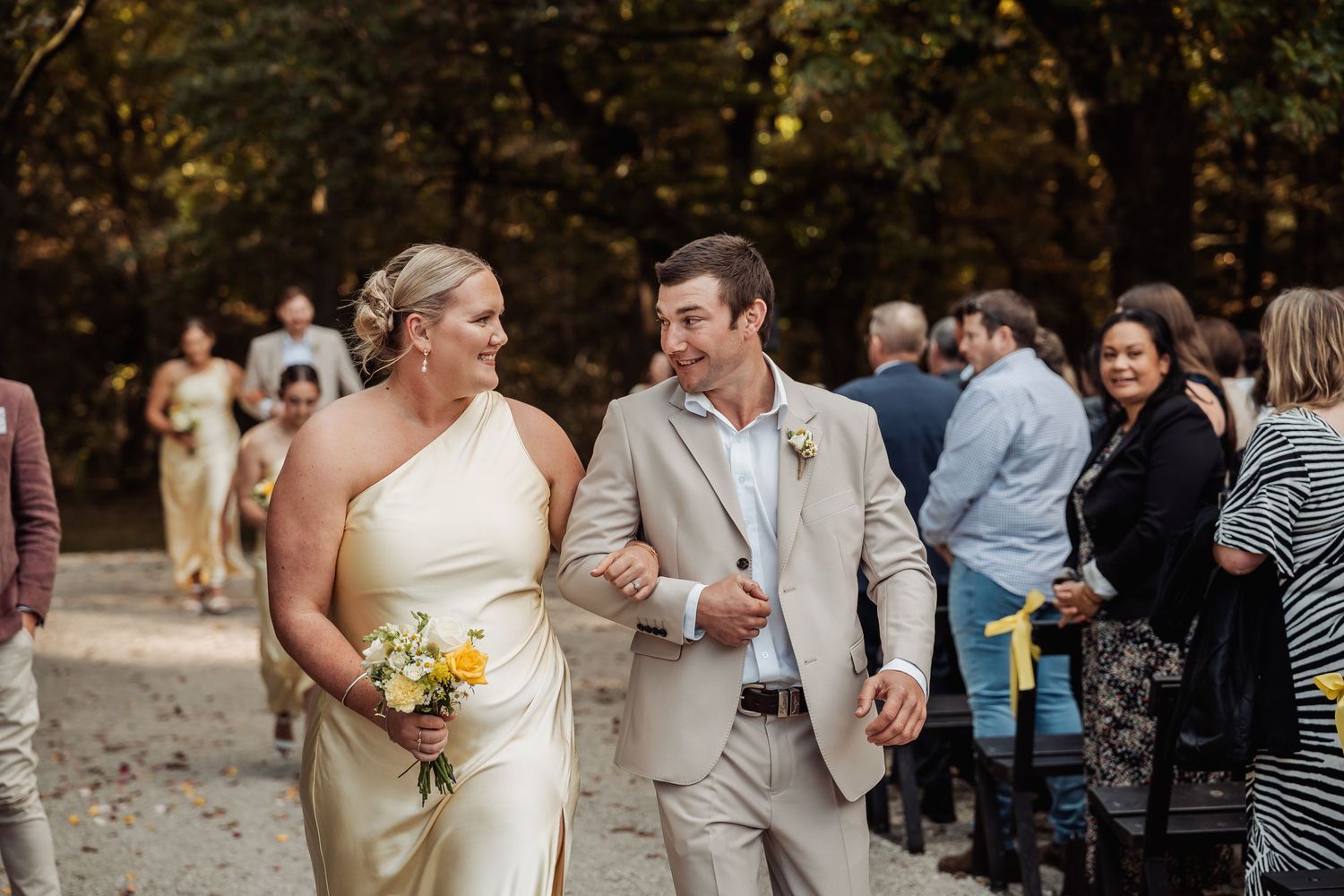 Bride in white one-shoulder gown walks with groomsman in tan suit after outdoor wedding ceremony.