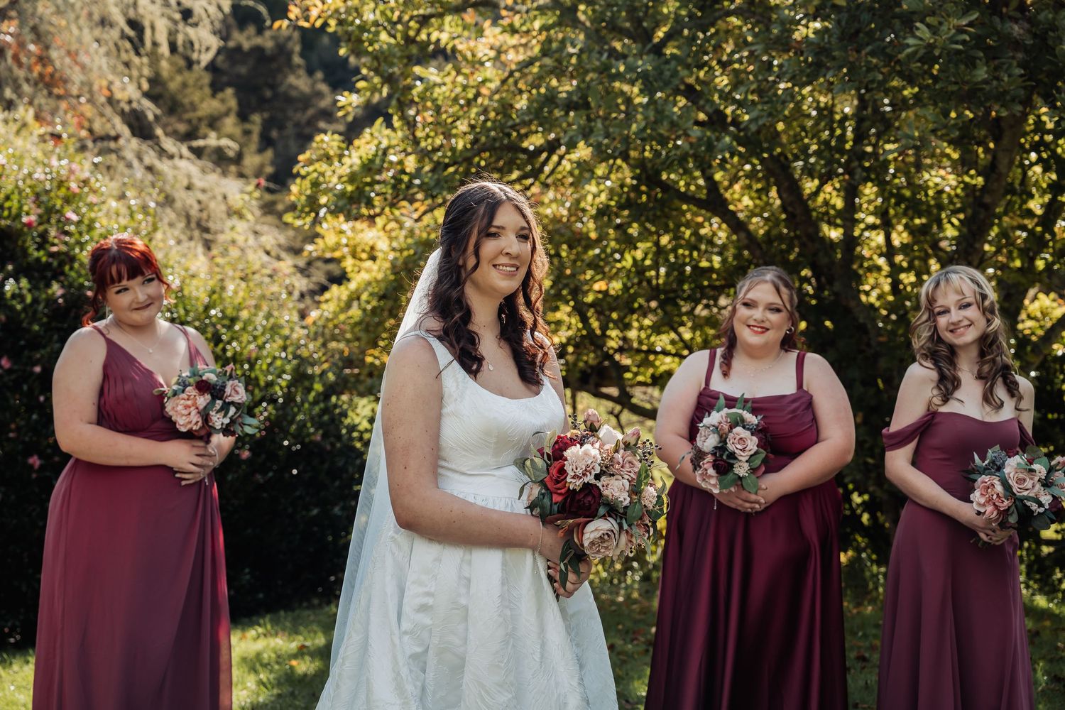 Bride in white gown holds bouquet with three bridesmaids in burgundy dresses outdoors among autumn trees.