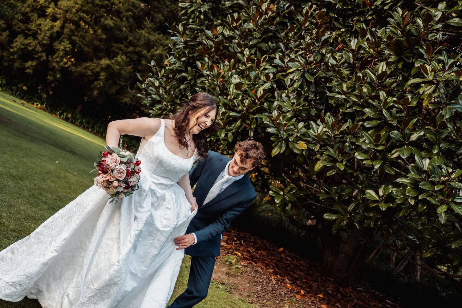 Bride in white gown and groom laughing together beside large green hedge outdoors.