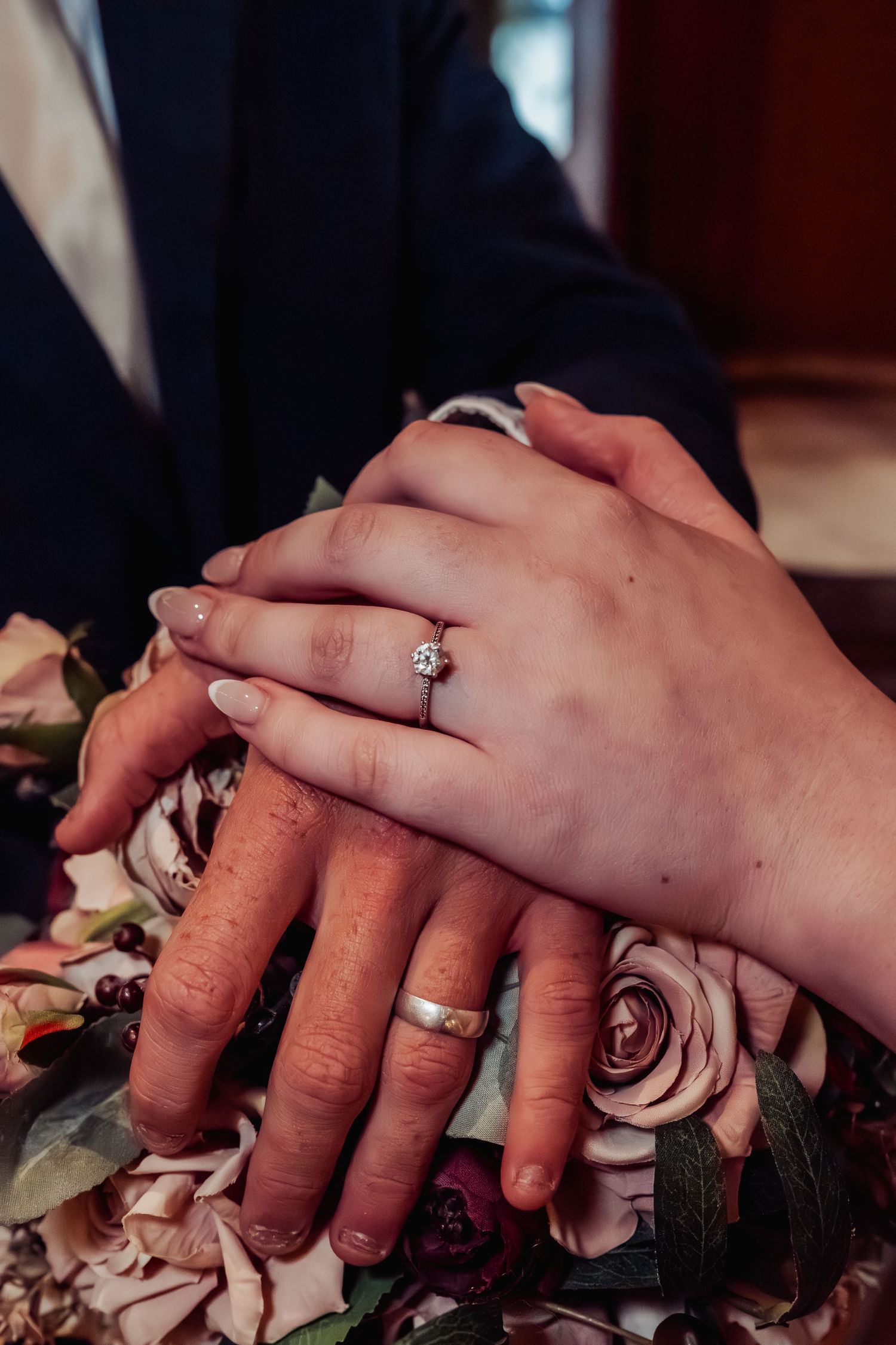 Close-up of newlyweds hands showing diamond engagement ring and gold wedding band over bouquet.