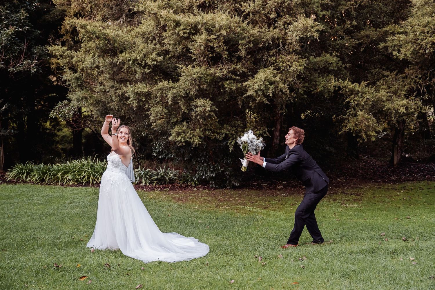 A playful wedding moment captured on grass as a suited figure kneels to present flowers.