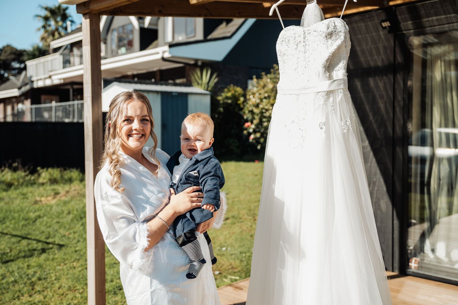 A white wedding gown hangs outside while someone stands nearby with a baby against a sunny backyard setting.