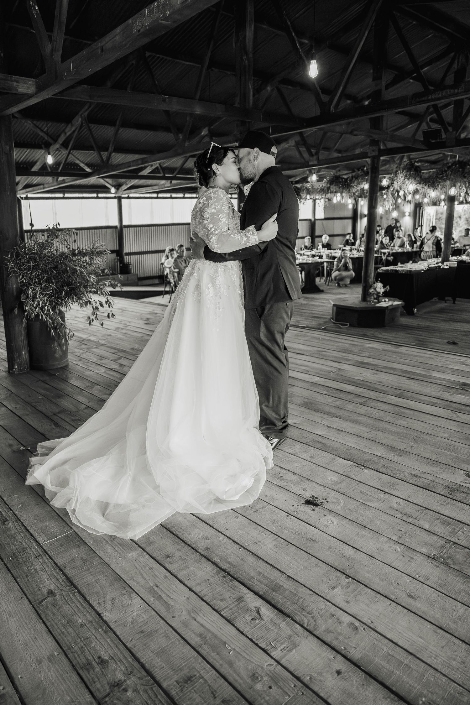 A black and white photograph captures an intimate wedding dance in a rustic barn venue with wooden floors and beams.