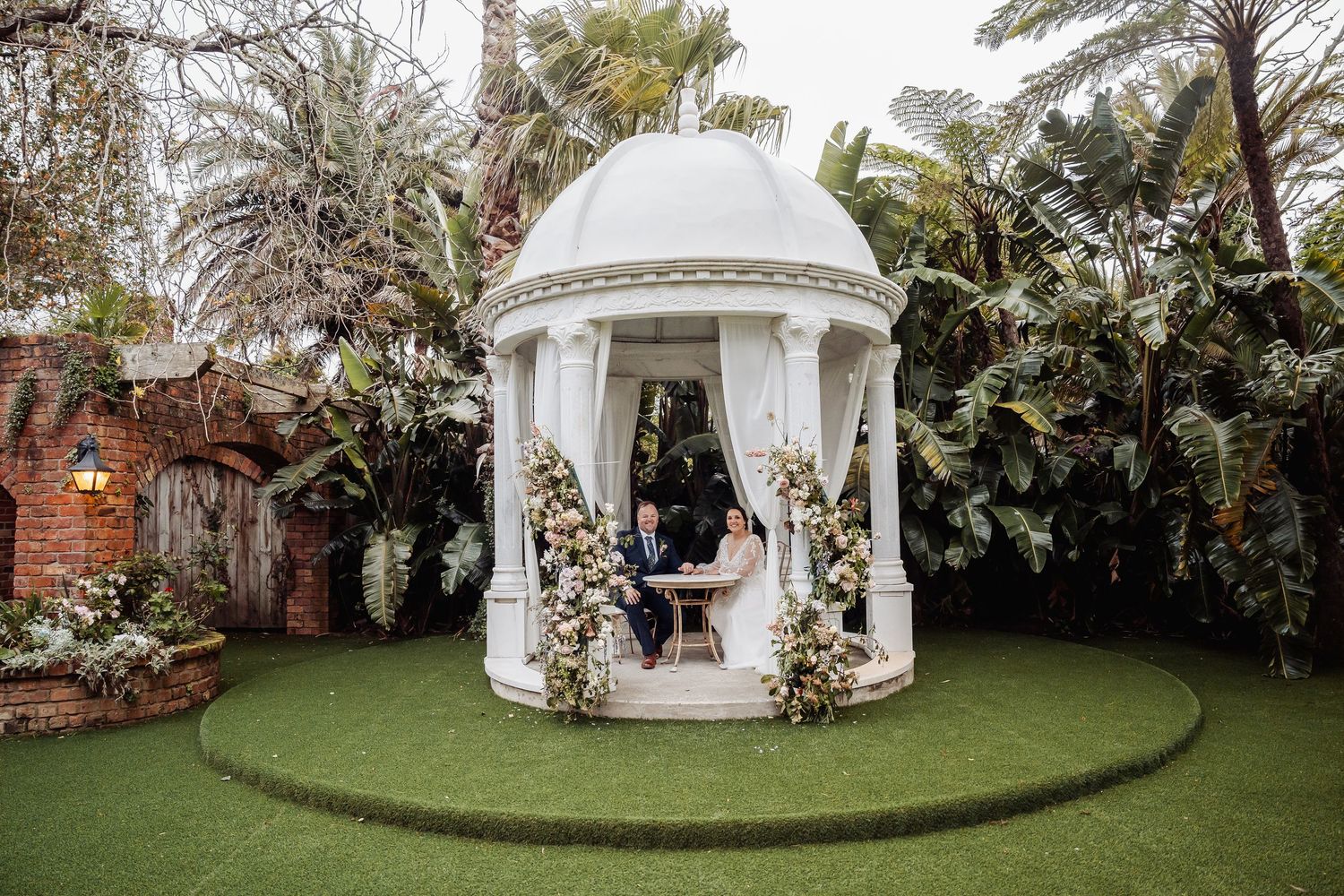 White garden gazebo surrounded by lush landscaping and palm trees.
