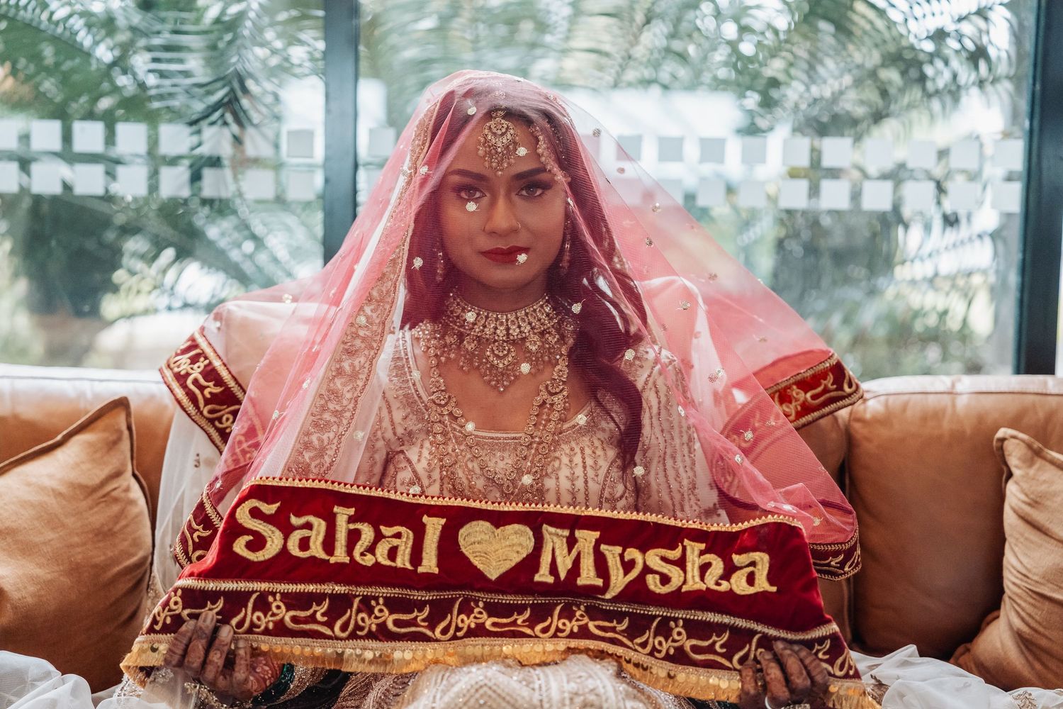 A traditional Indian bride in a pink embellished lehenga and veil holds a sign reading 'Sabal loves Mysha'.