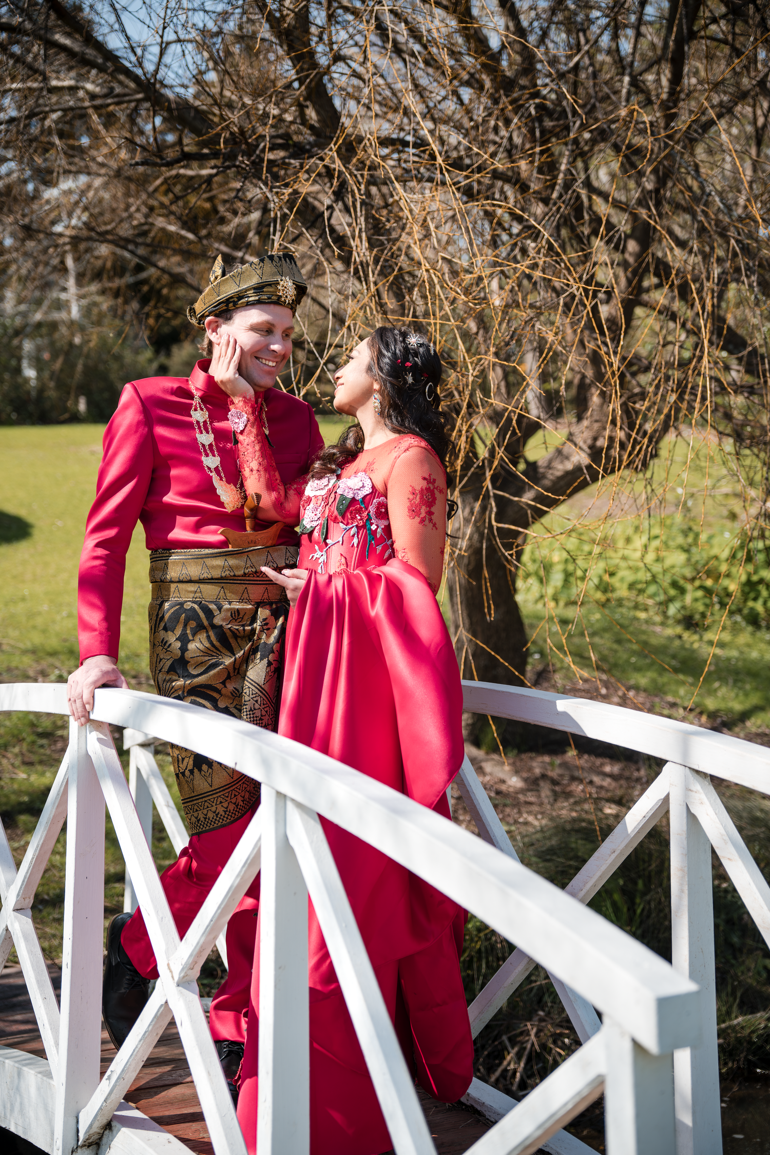 A couple in matching red formal attire share a romantic moment on a white bridge.