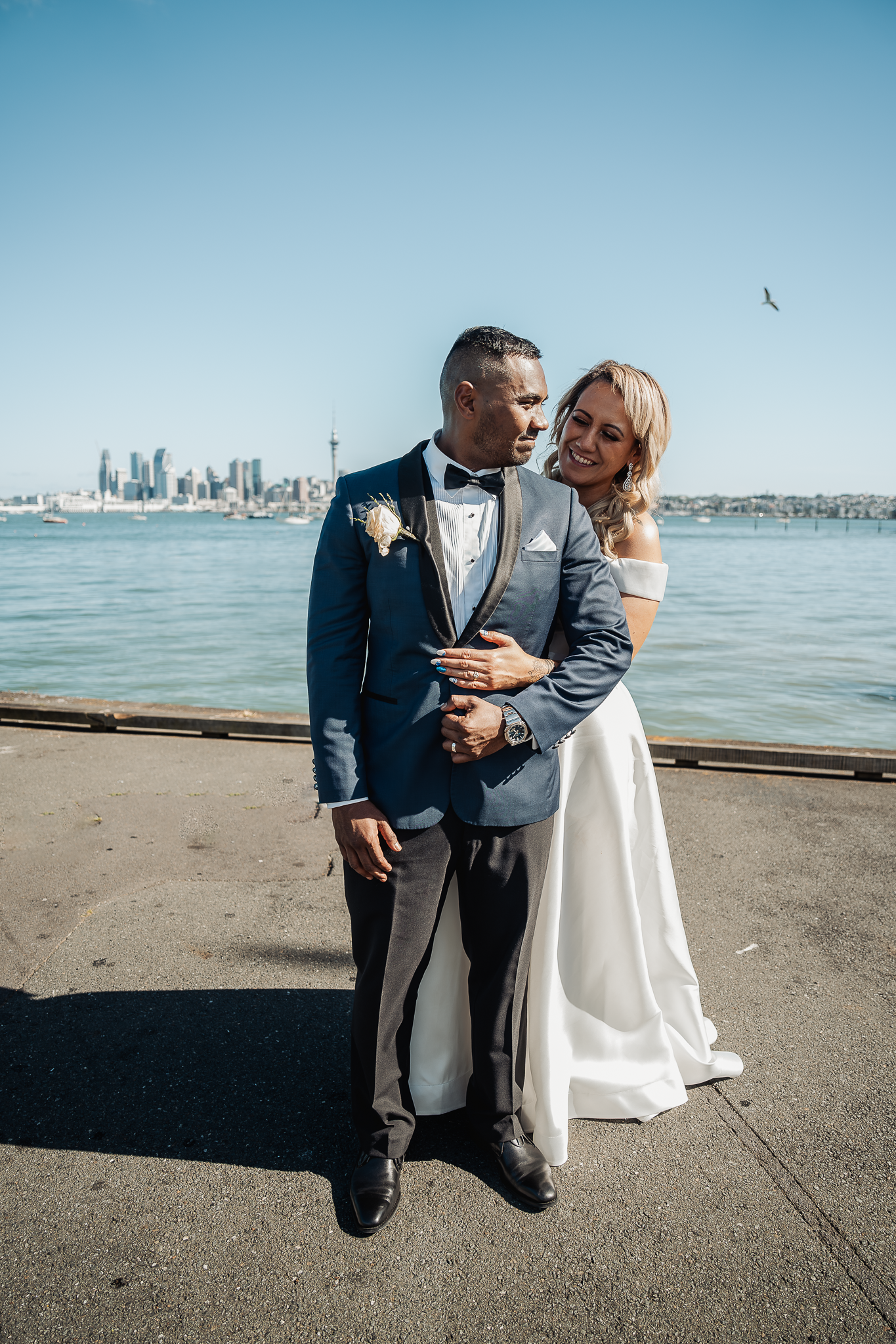 Wedding couple poses on waterfront pier with city skyline in background.