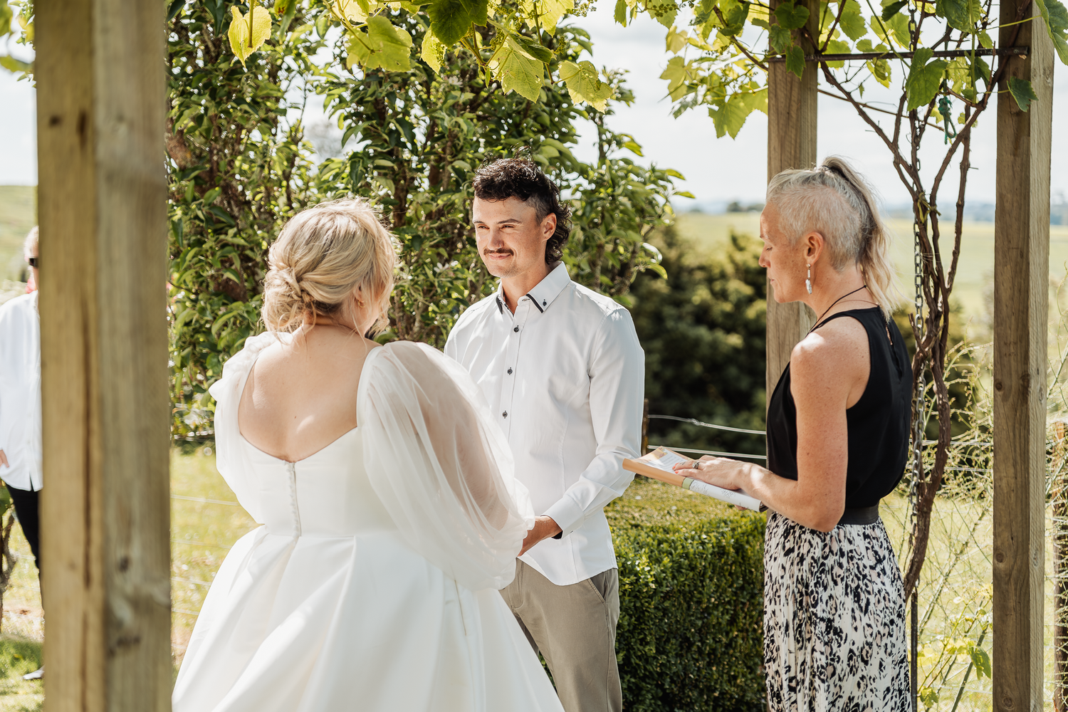 A wedding ceremony takes place under a wooden pergola surrounded by lush greenery.