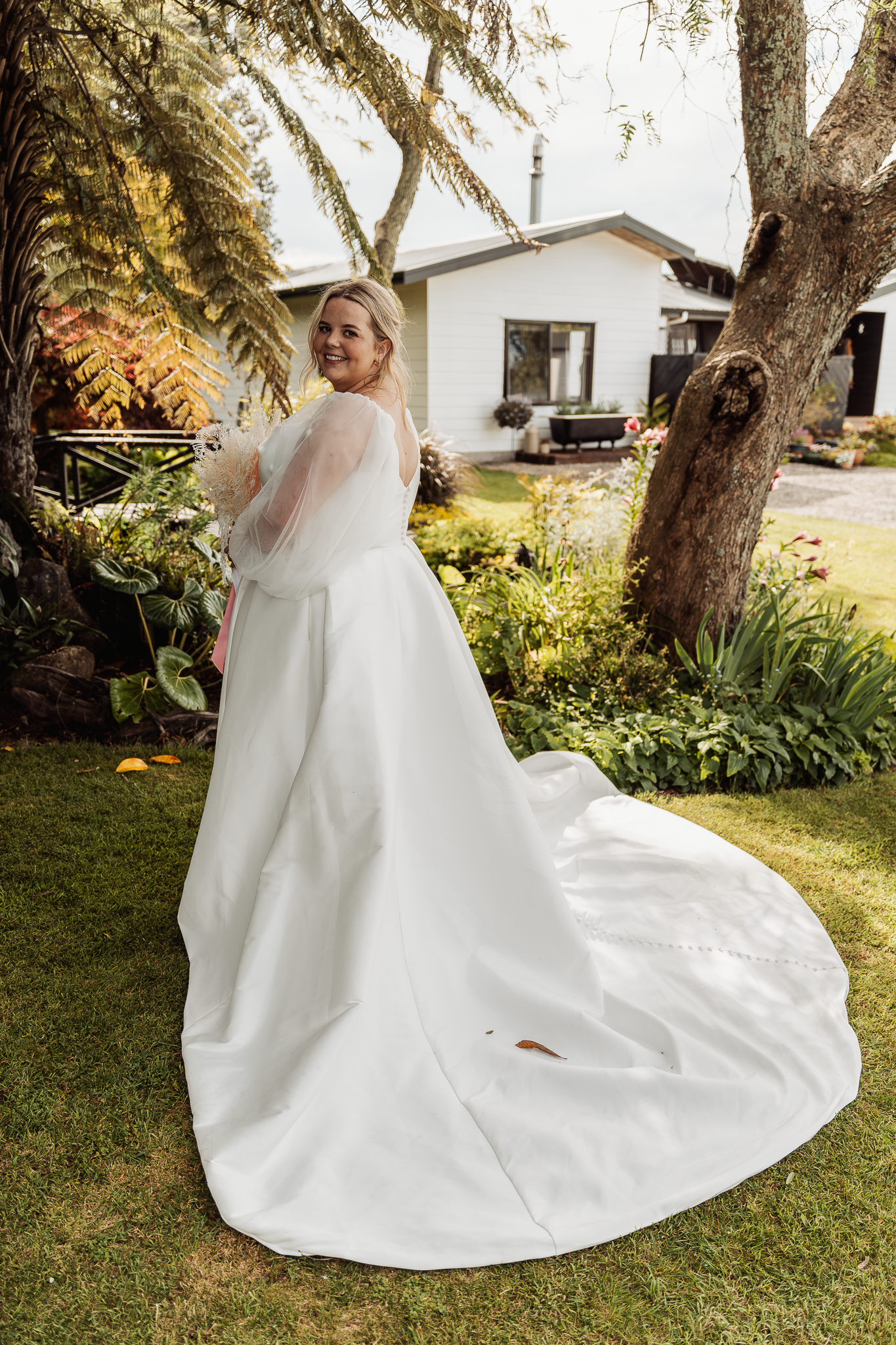 Elegant bride in white gown with dramatic train poses in a garden setting with house in background.