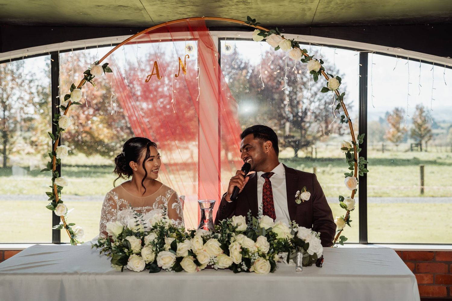 Bride and groom smiling at sweetheart table with white floral centerpiece and circular floral arch.