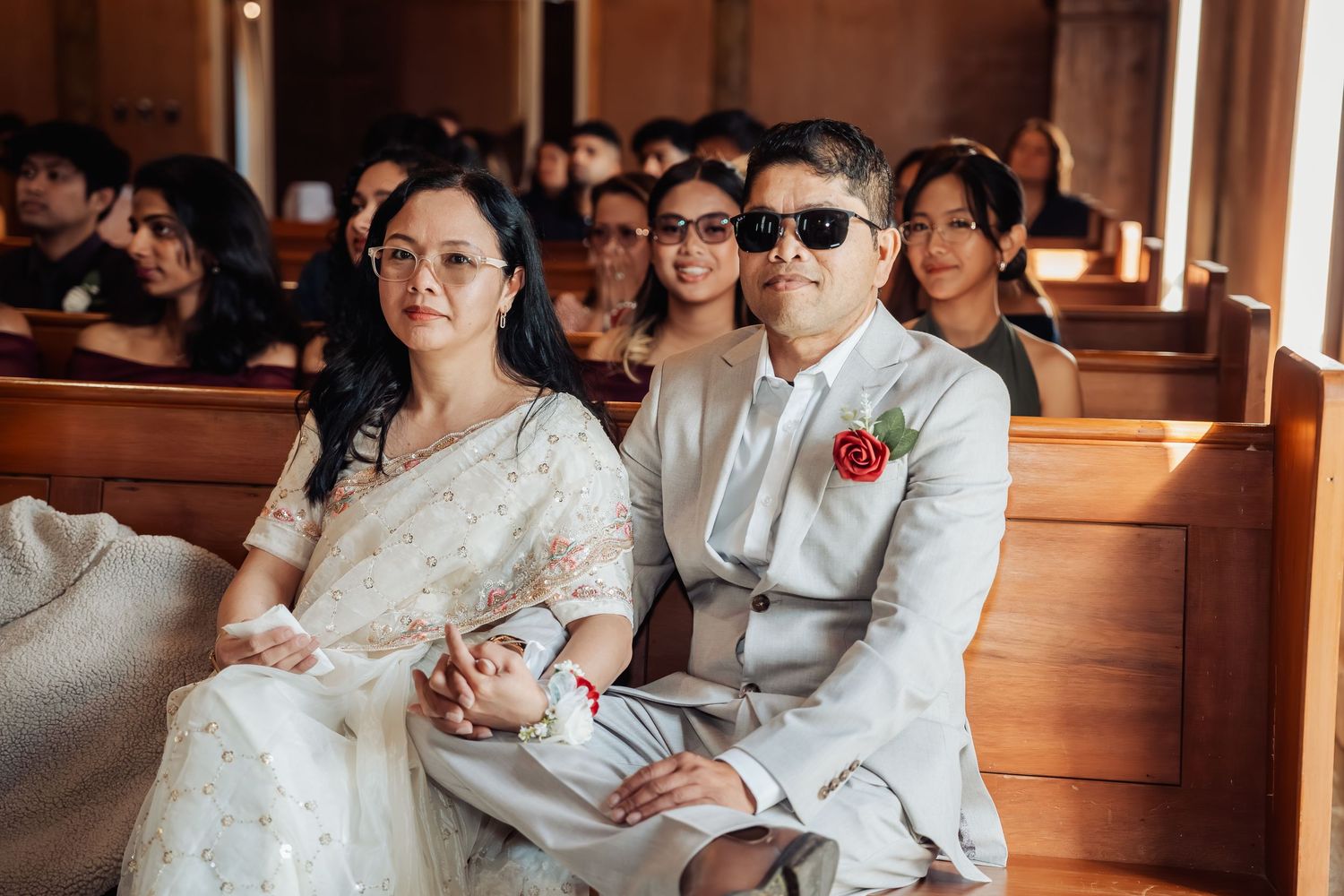 Wedding guests in traditional attire sit in wooden church pews during ceremony.