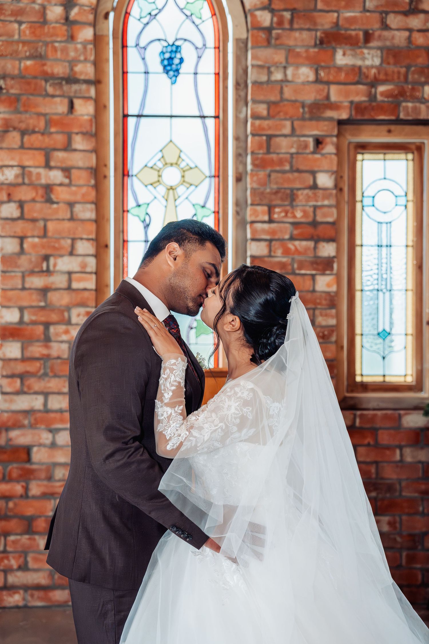 Bride and groom sharing a romantic moment in front of a stained glass window in a brick church.