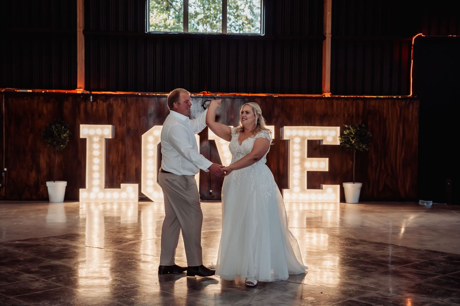 Bride and groom share first dance beneath illuminated LOVE letters at rustic barn wedding reception.