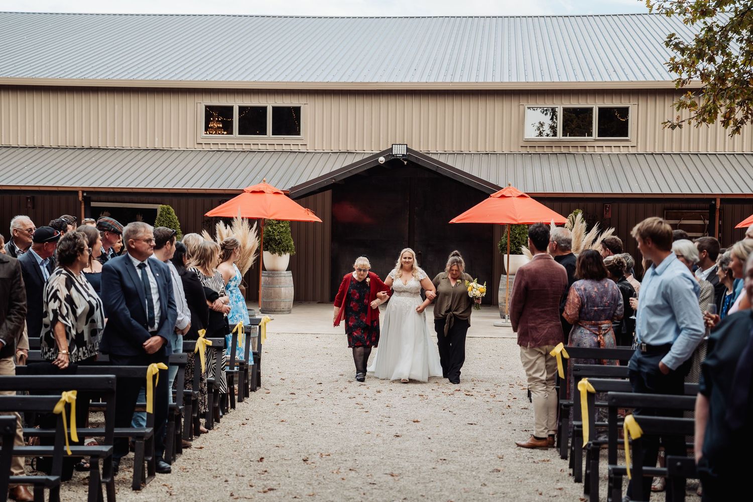 Bride walking down outdoor aisle at rustic barn wedding ceremony with guests seated.