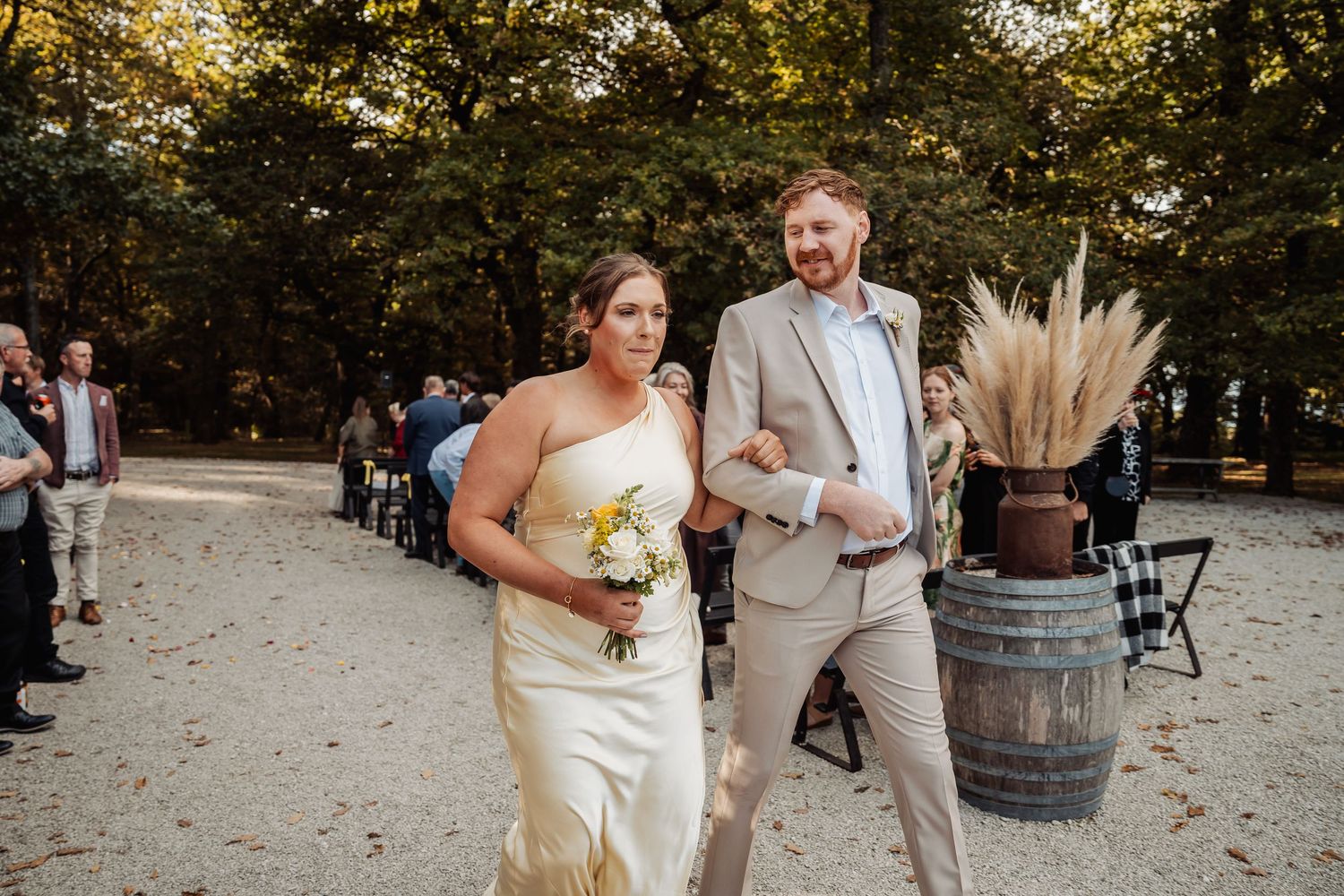 Bride and groom walk down sandy outdoor wedding aisle surrounded by autumn trees and guests.