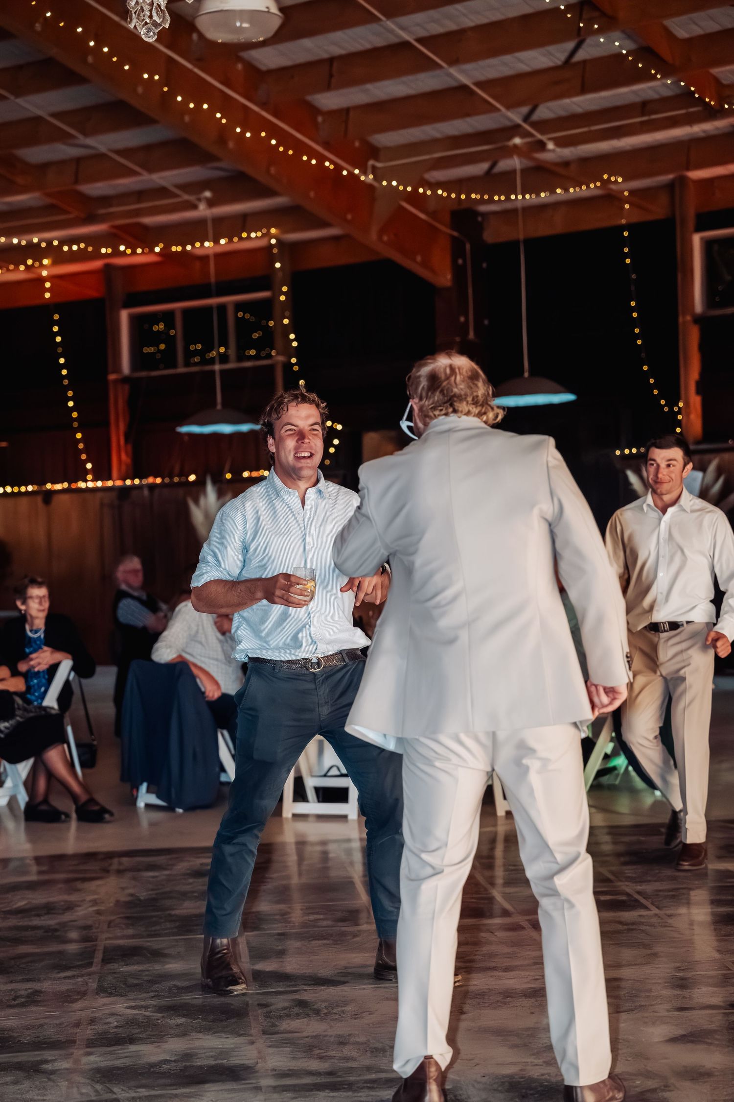 Two men dance joyfully together on decorated barn dance floor at wedding reception.