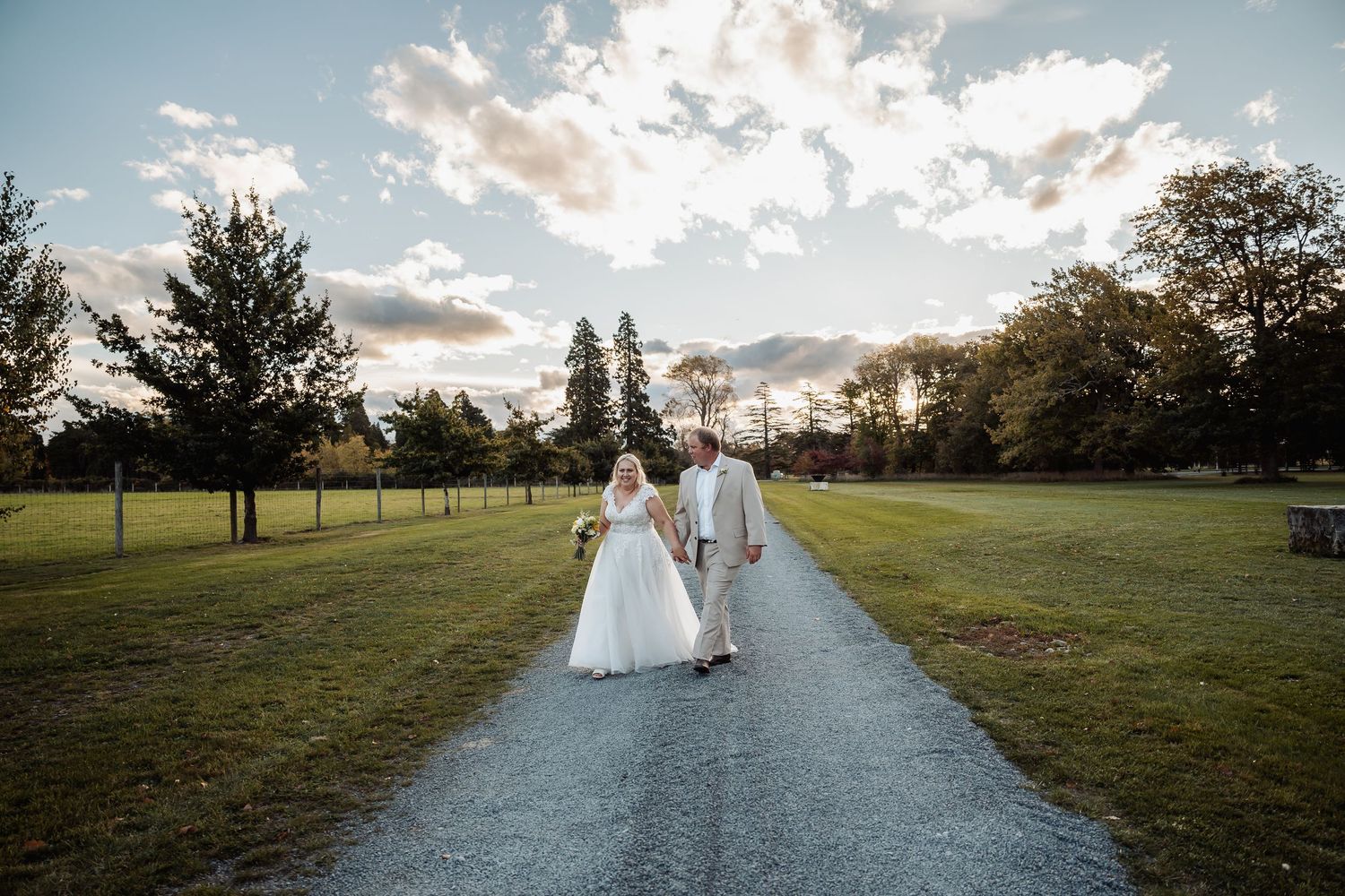 Bride and groom walk hand in hand down gravel path through lush green park at golden hour sunset.