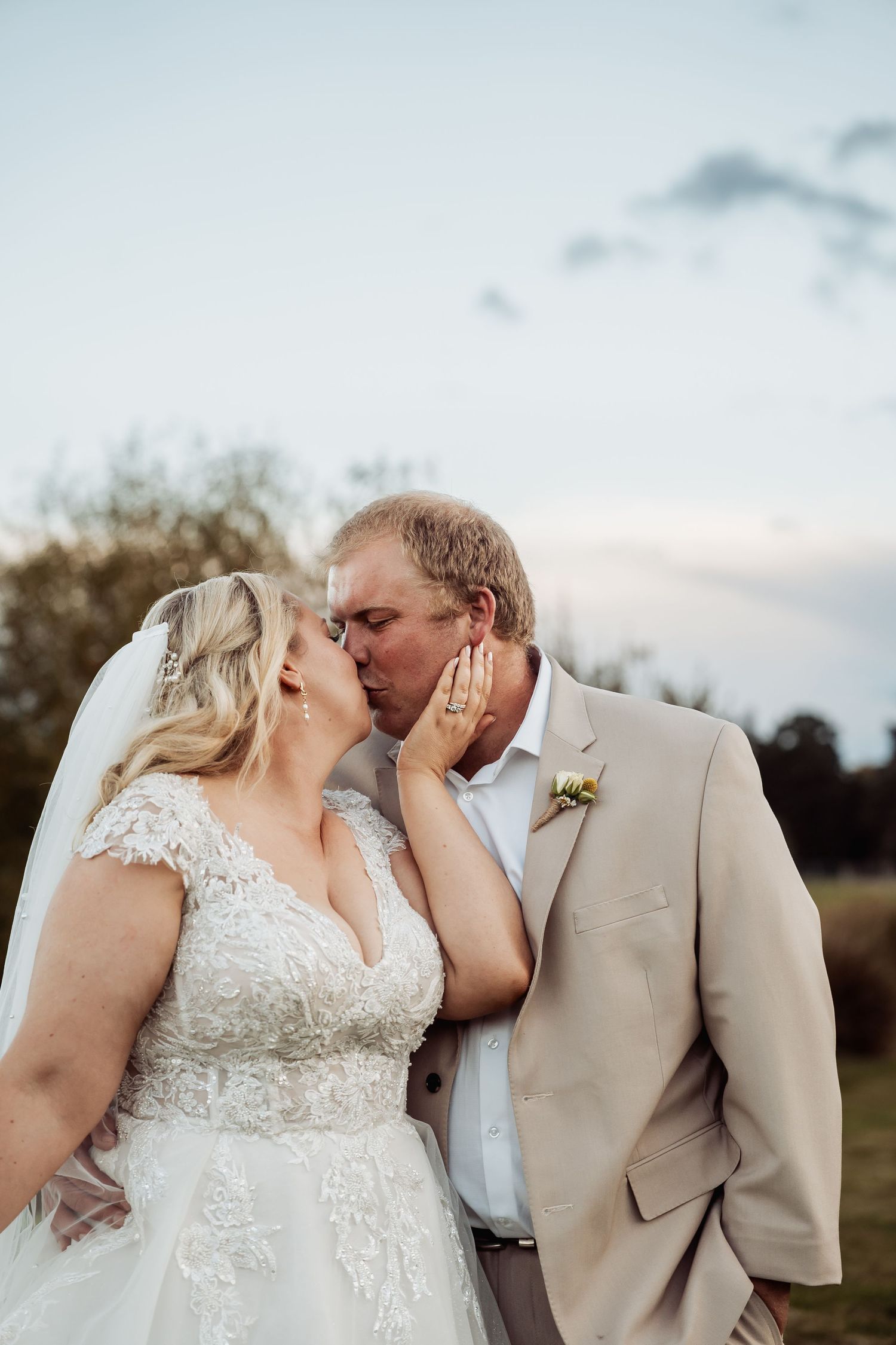 Bride and groom share romantic kiss at sunset, bride in lace gown with veil, groom in tan suit.