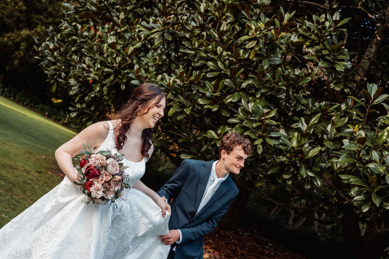 Bride in white ball gown holds bouquet while groom in navy suit smiles beside lush greenery.