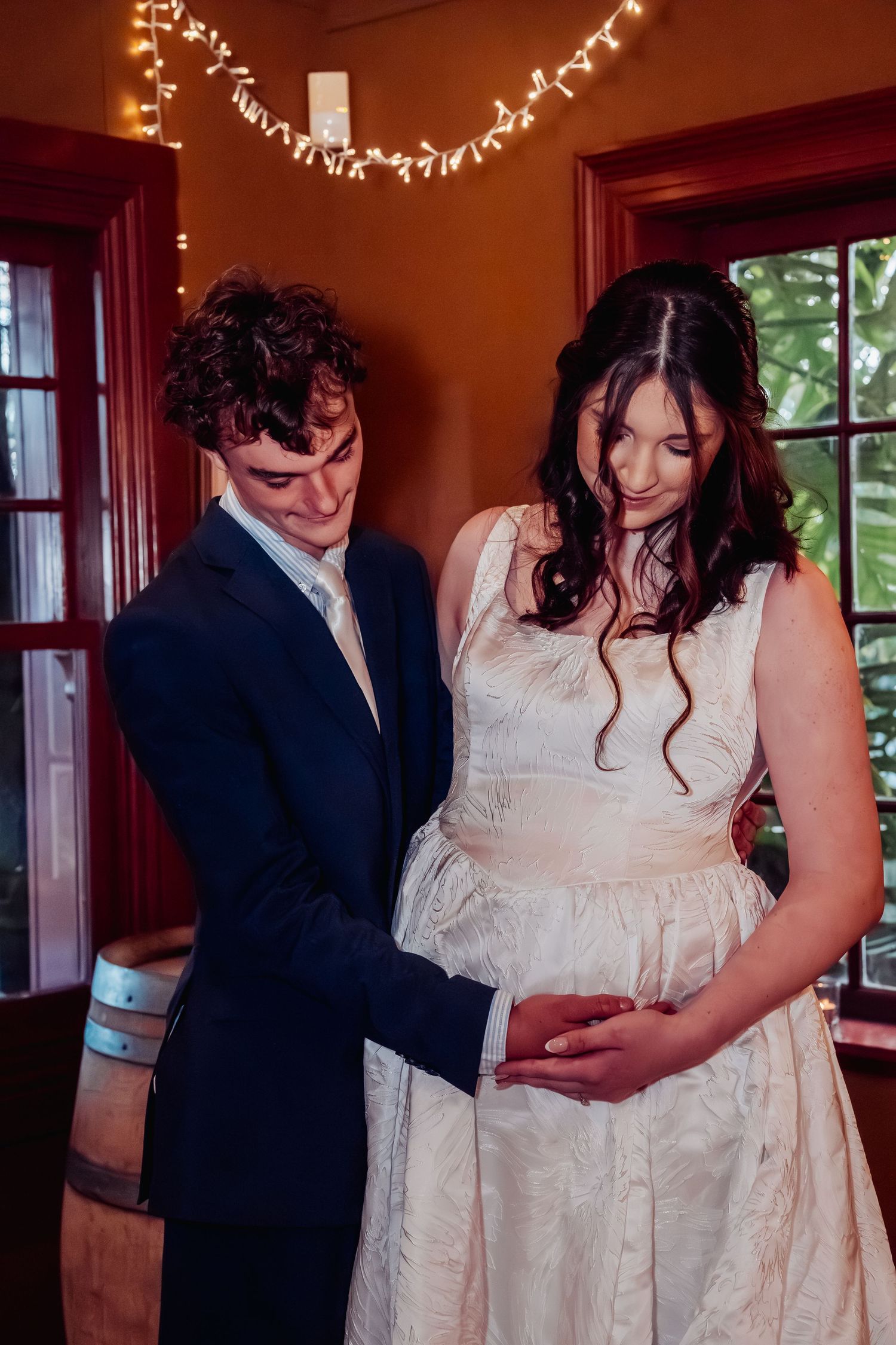 Bride and groom tenderly cradle baby bump together in warmly lit wedding venue.