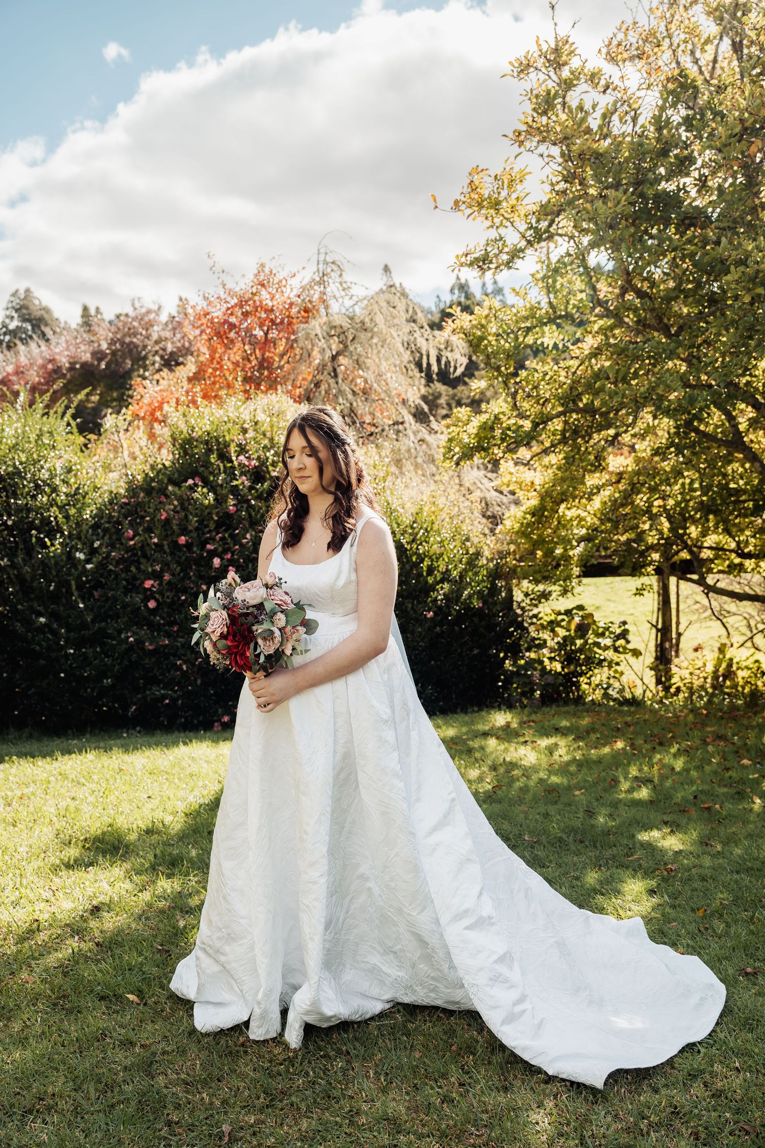 Bride in white gown holding bouquet standing in lush garden on sunny wedding day.
