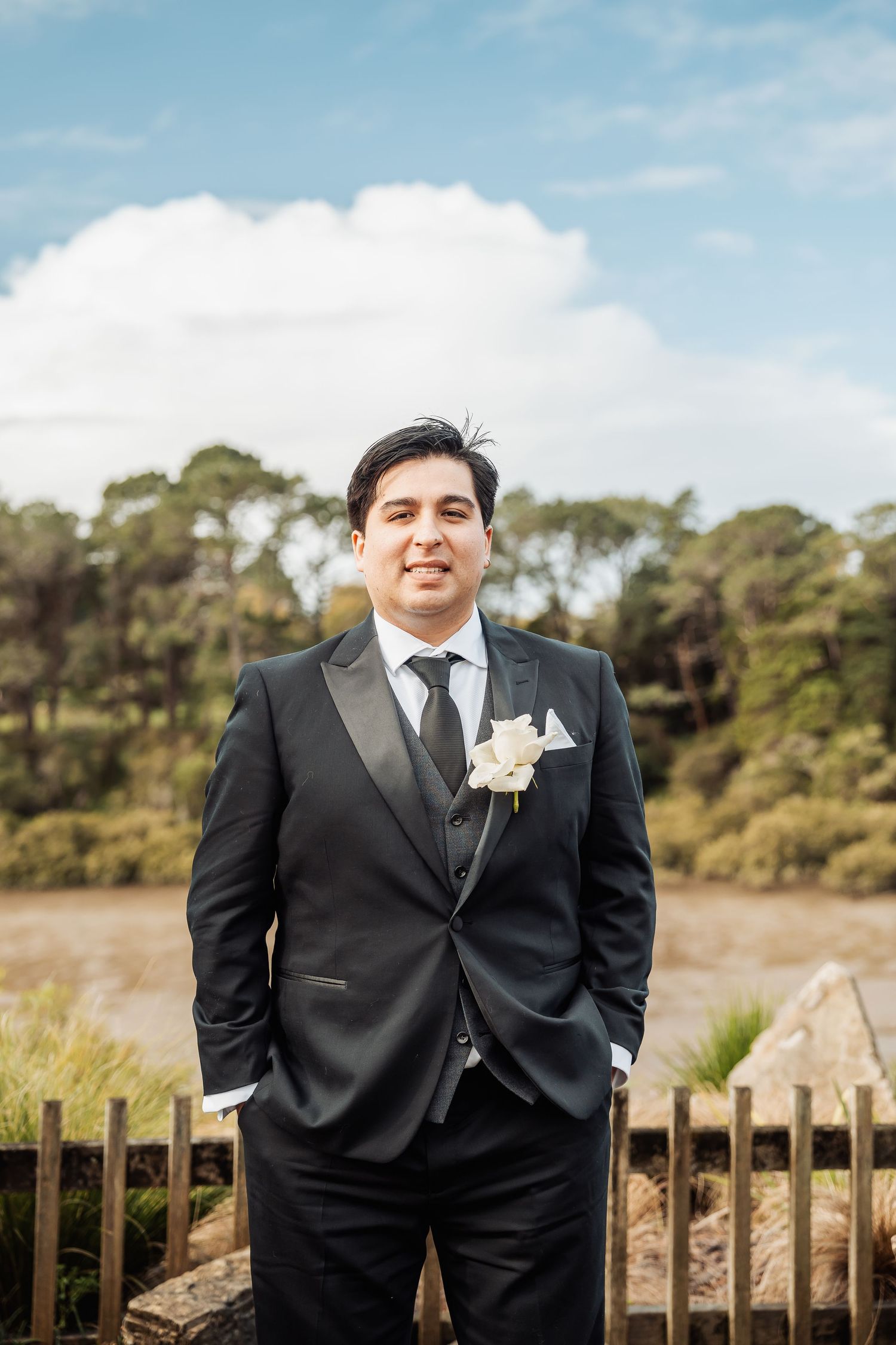 A groom in a black tuxedo poses outdoors with a white boutonniere against a scenic natural background.