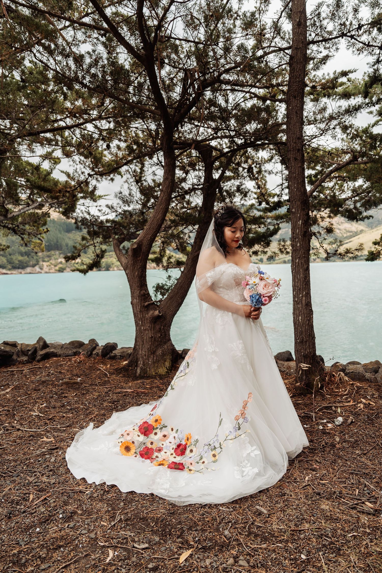 Bride in floral embroidered wedding gown holding colorful bouquet standing among pine trees by lake.