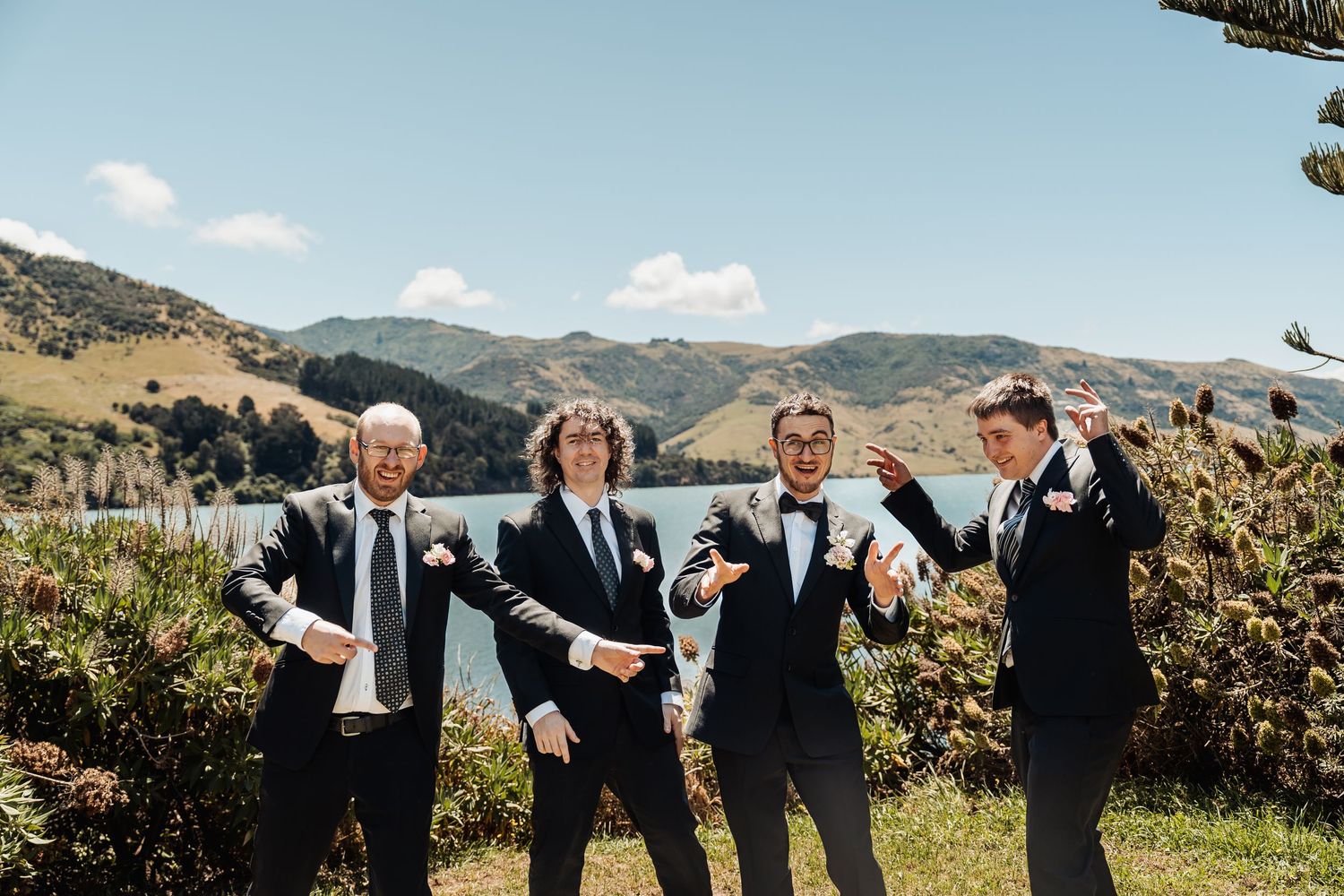 Four groomsmen in black suits posing playfully by a scenic lake with green hills in New Zealand.