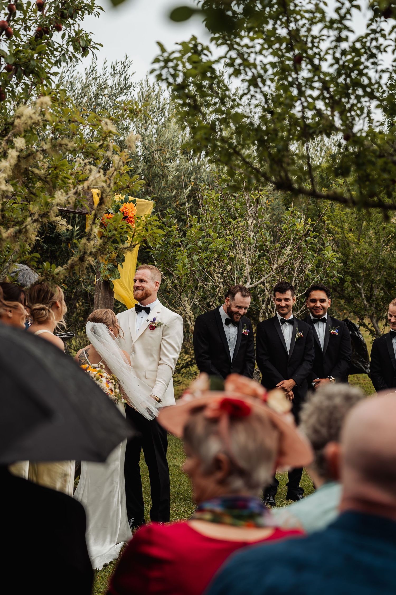 Groom in white tuxedo laughing with bride during outdoor garden wedding ceremony with guests.