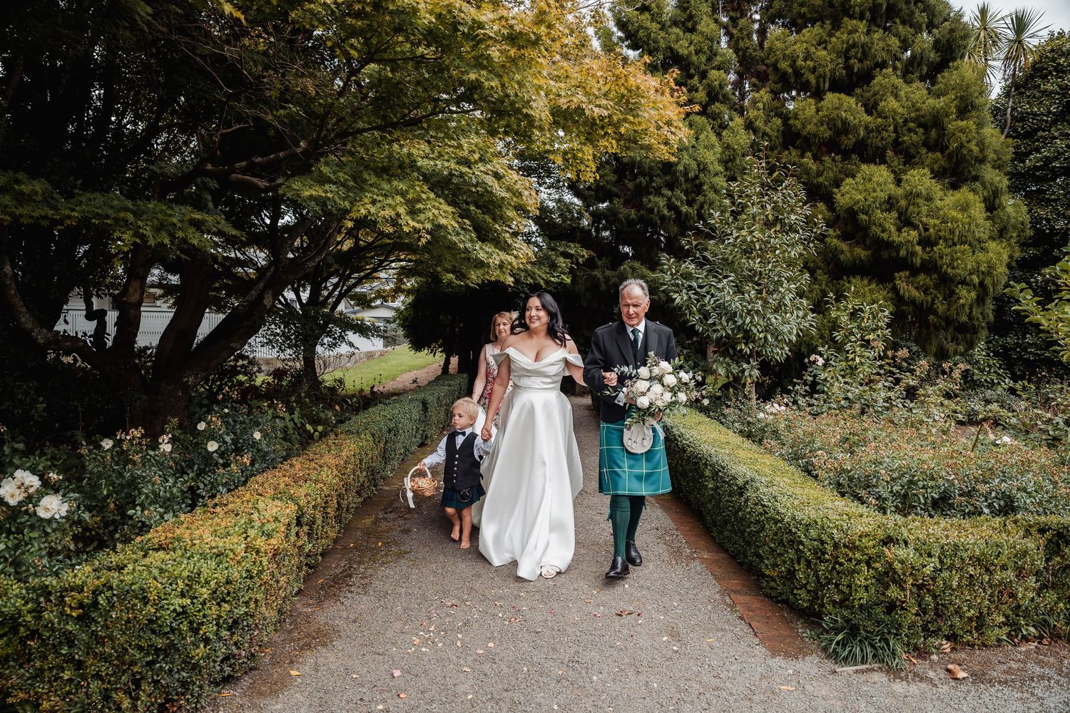 Bride in white gown walks garden path with man in Scottish kilt and child at outdoor wedding ceremony.