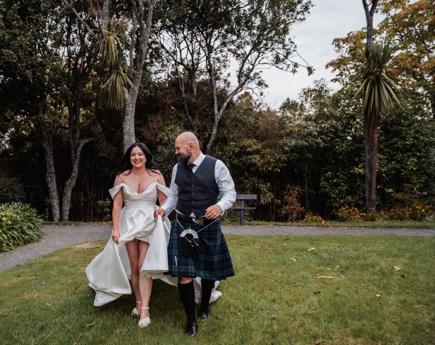 Bride lifting dress while walking with groom in Scottish kilt through lush garden grounds.