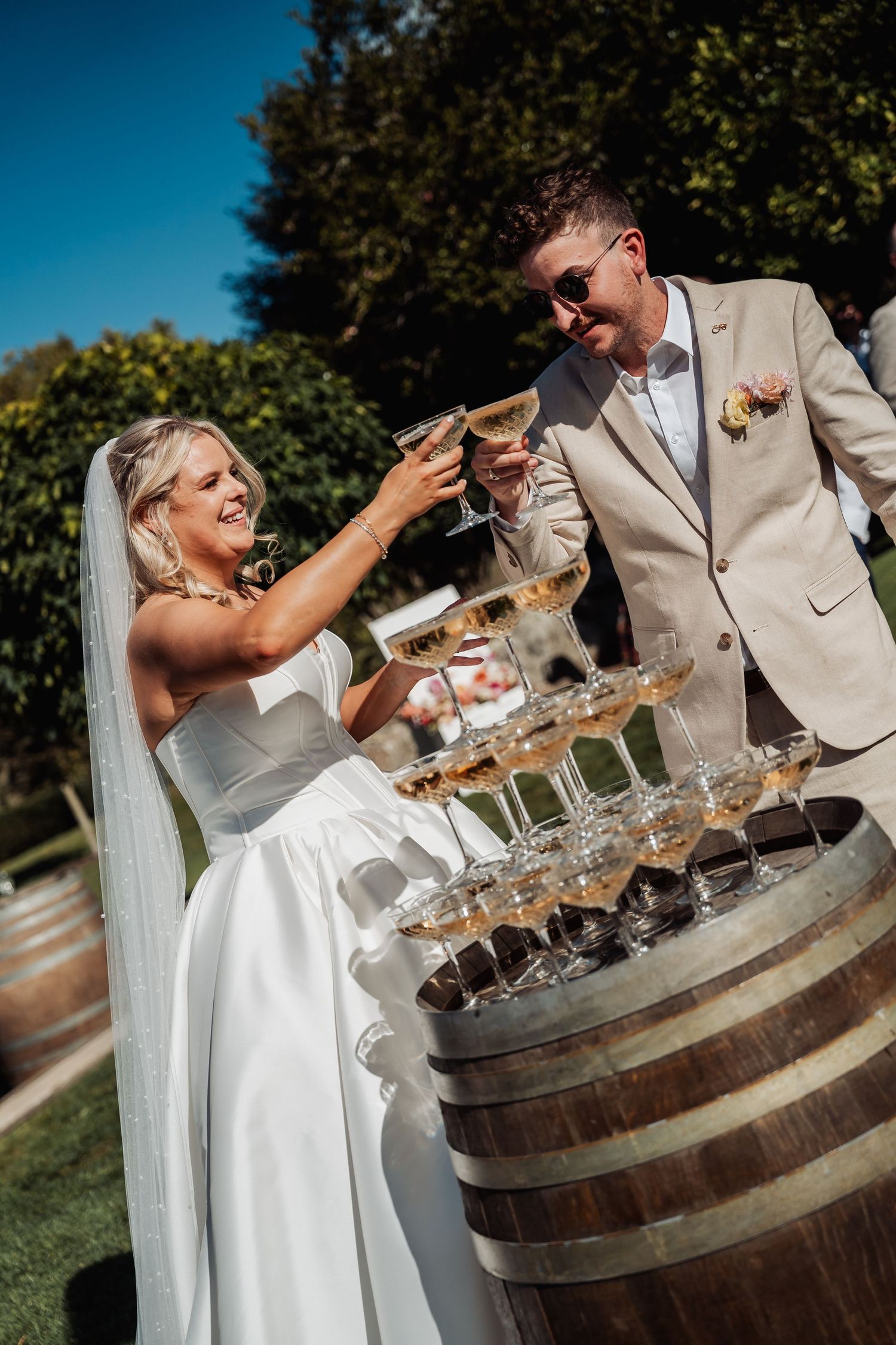 Bride and groom toasting champagne glasses over a pyramid tower on a wooden barrel at an outdoor wedding.