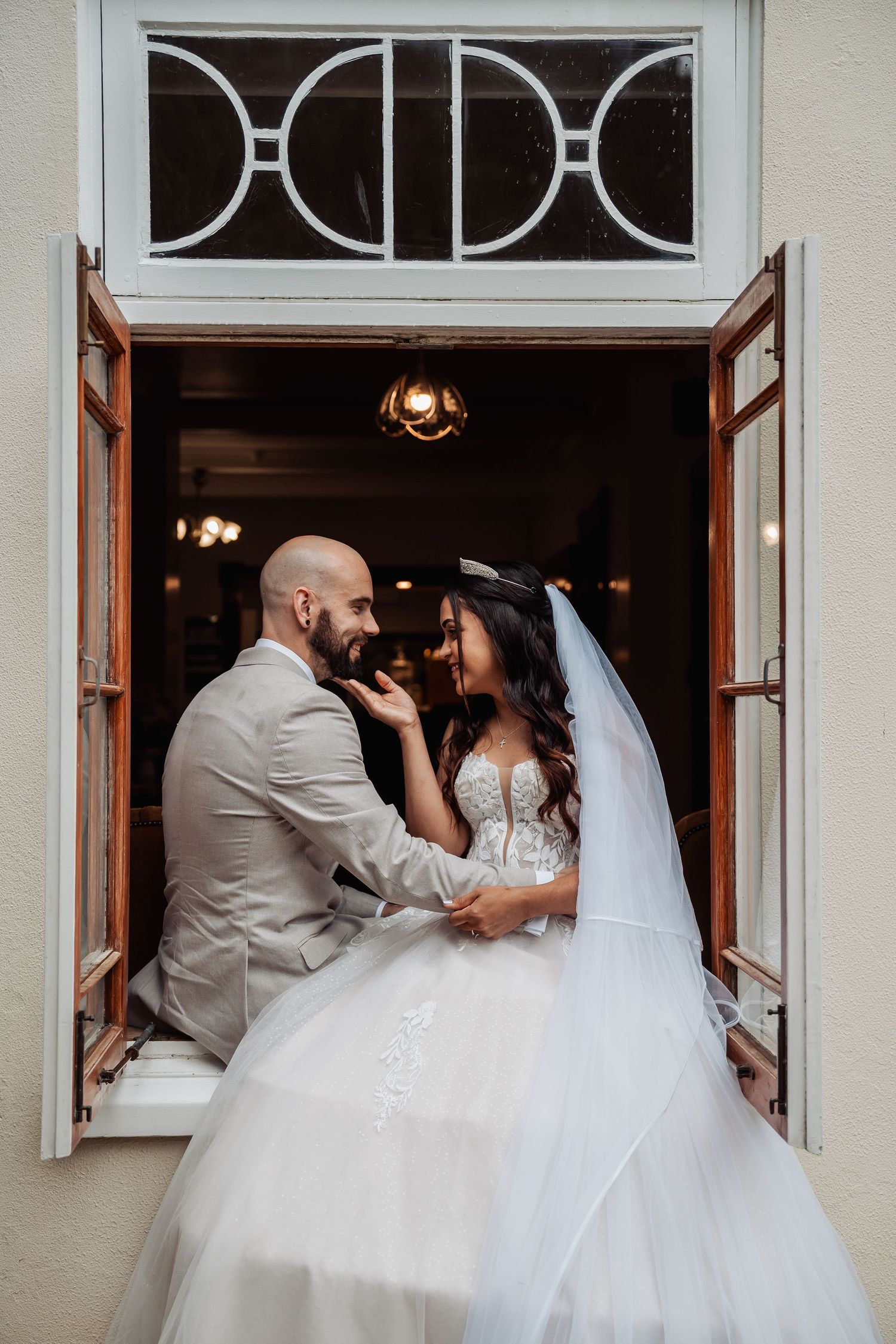 Bride and groom share romantic moment framed by vintage art deco window with warm interior lighting.