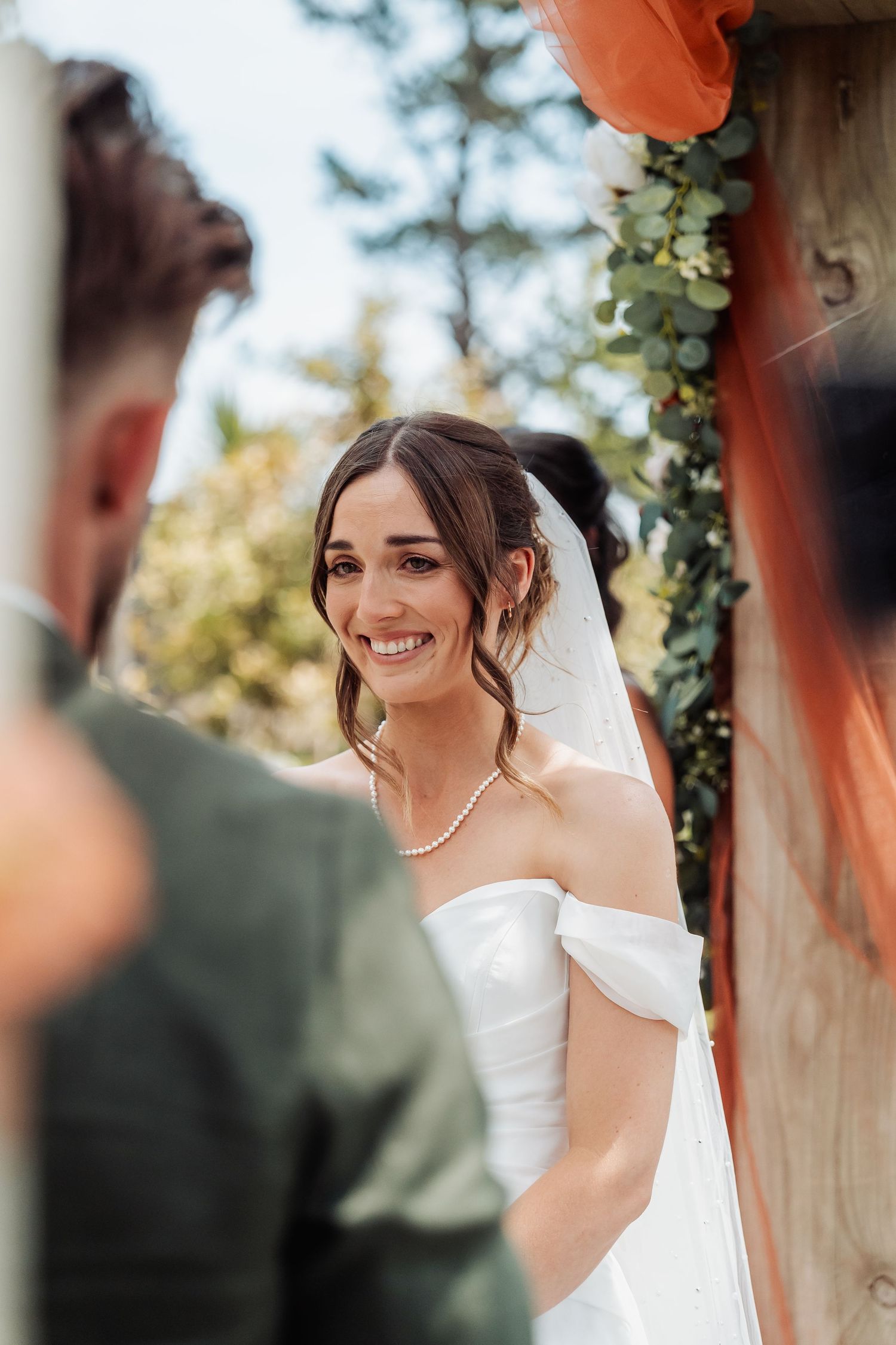 Radiant bride in off-shoulder white gown and veil smiling at groom during outdoor wedding ceremony.