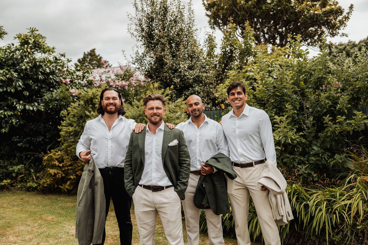 Four groomsmen in smart casual attire smile together in a lush garden setting.