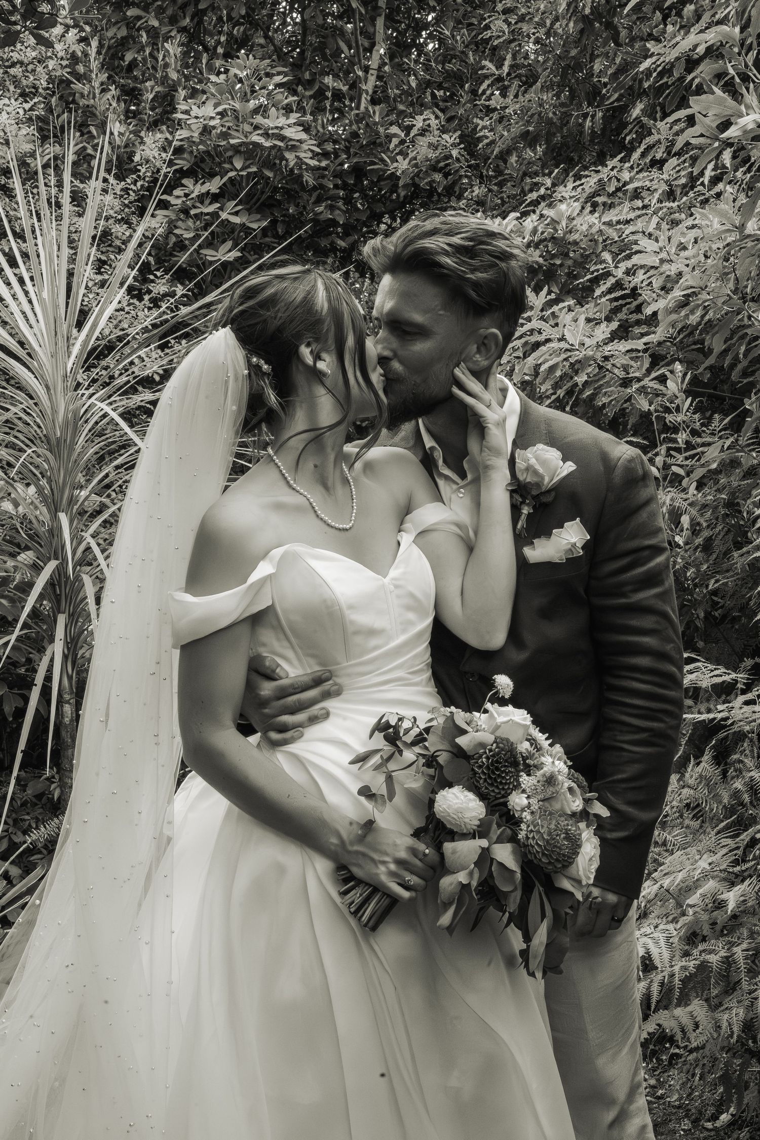 Black and white photo of bride and groom kissing in garden, bride holding floral bouquet.