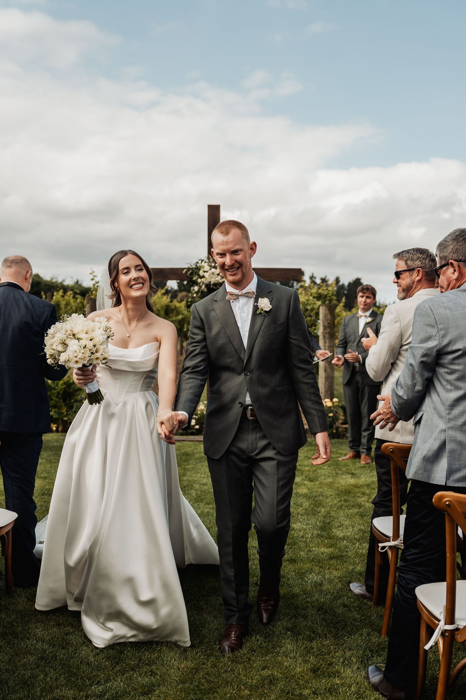Newlyweds walk joyfully down the aisle after outdoor wedding ceremony with guests applauding around them.