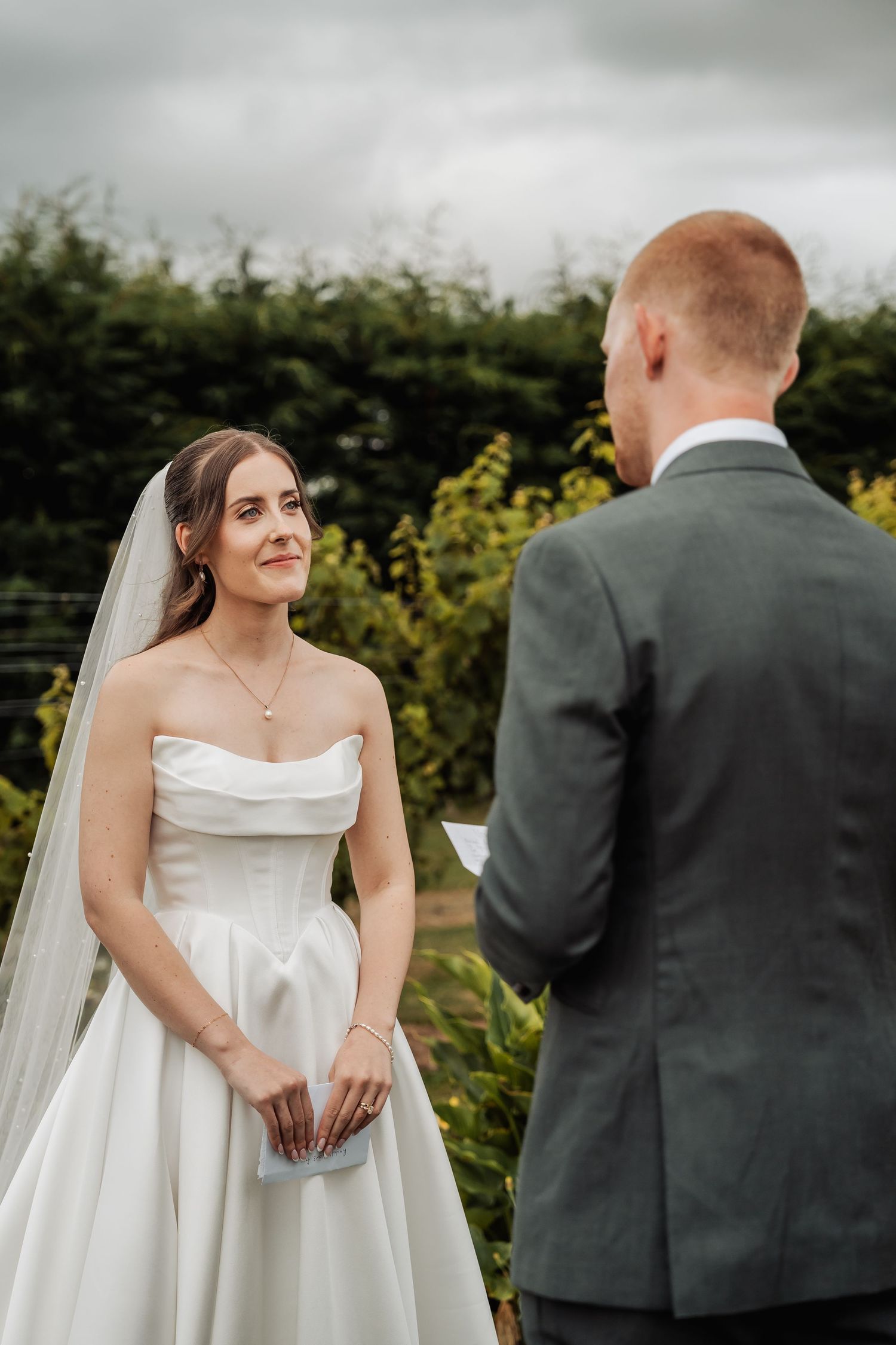 Bride smiles lovingly at groom during outdoor wedding ceremony vows surrounded by green hedges.